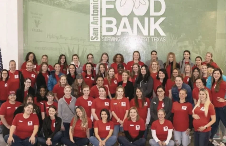 Group of women at a food bank event in San Antonio, Texas, with many wearing red shirts and posing for a group photo in front of a large sign that reads 'Food Bank.'
