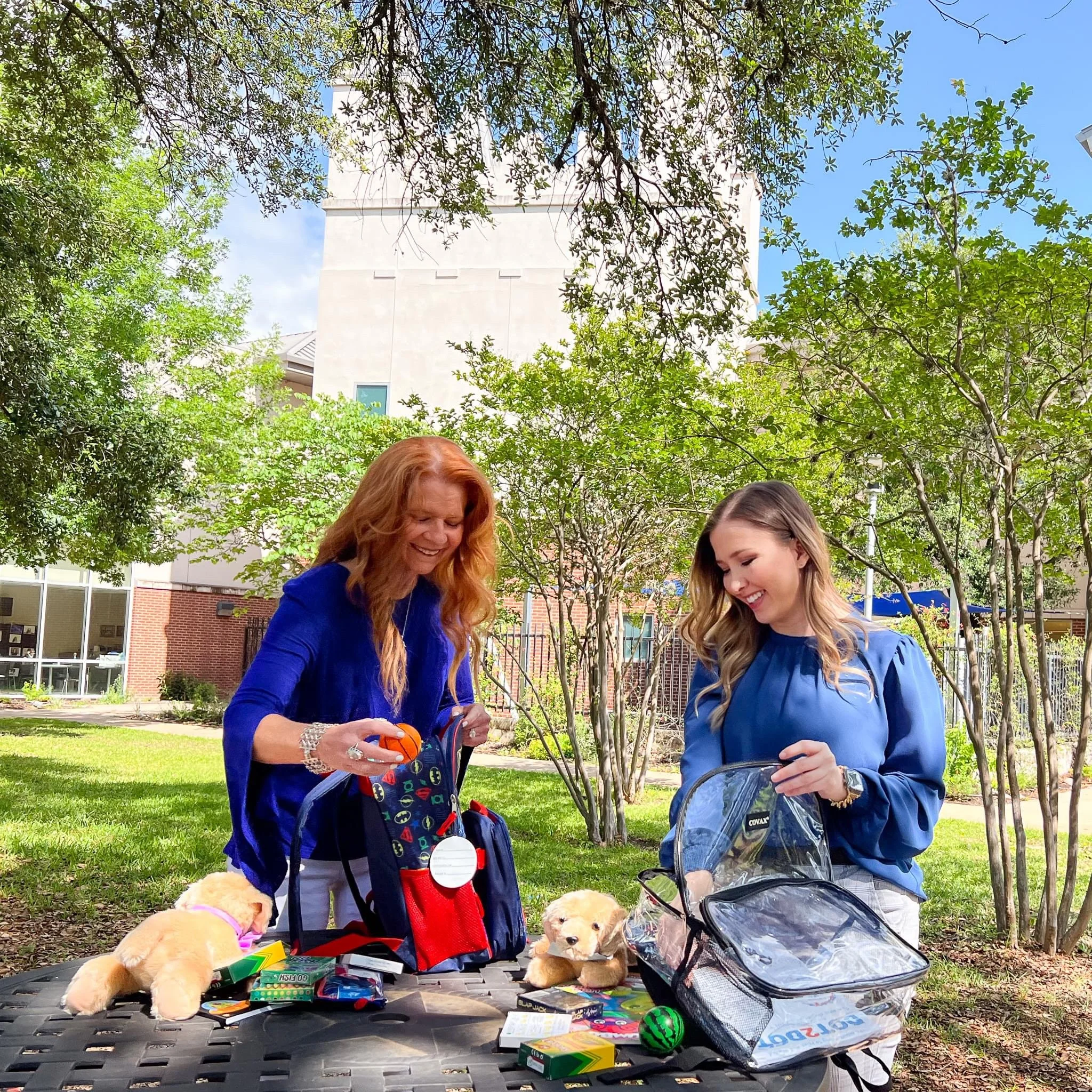 Two women packing school supplies into backpacks outdoors on a sunny day, with trees, grass, and a building in the background.