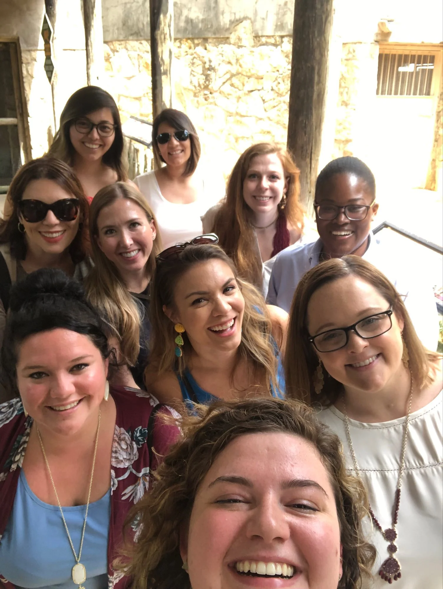 Group of ten smiling women taking a group selfie outdoors in front of a stone wall.