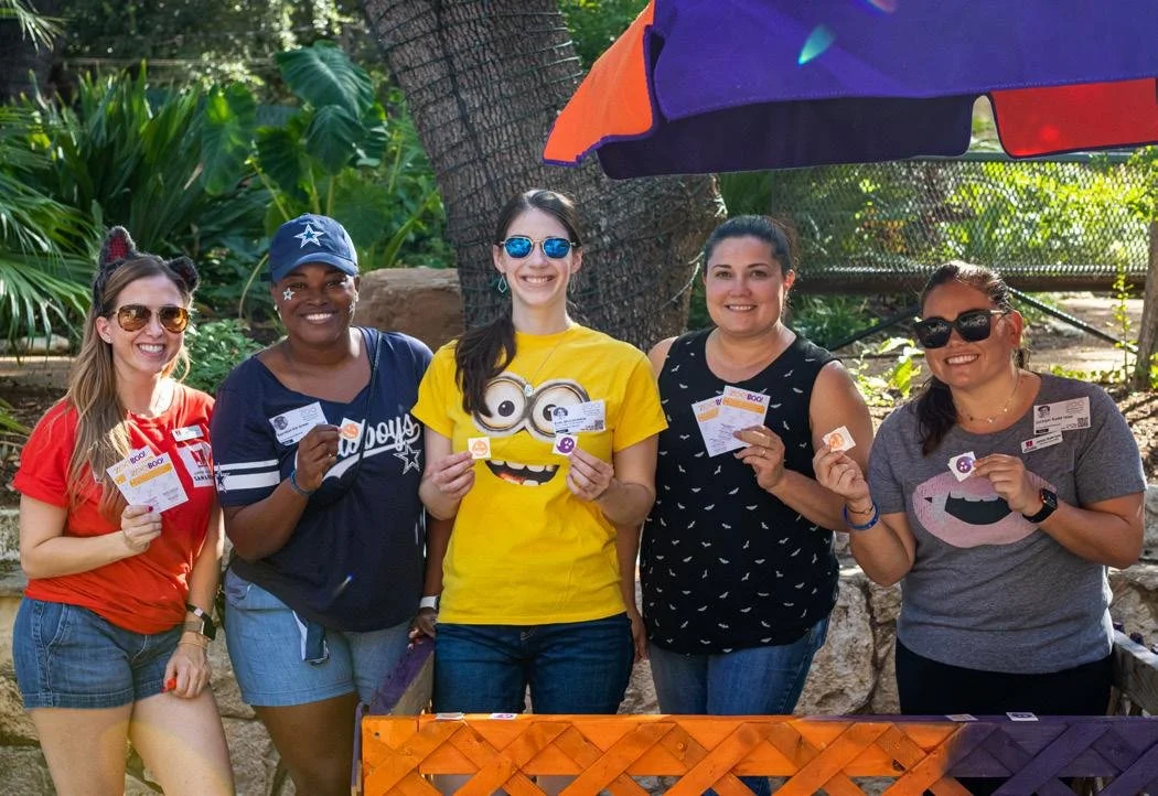 Five women standing outdoors under an umbrella, smiling and holding tickets or passes, in a lush, green setting.