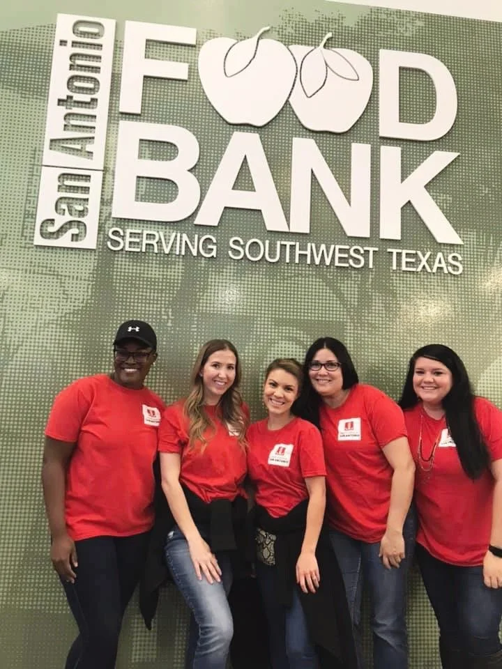 Five women wearing red shirts standing in front of a green Wall with a sign that says 'San Antonio Food Bank Serving Southwest Texas,' at a food bank event or location.