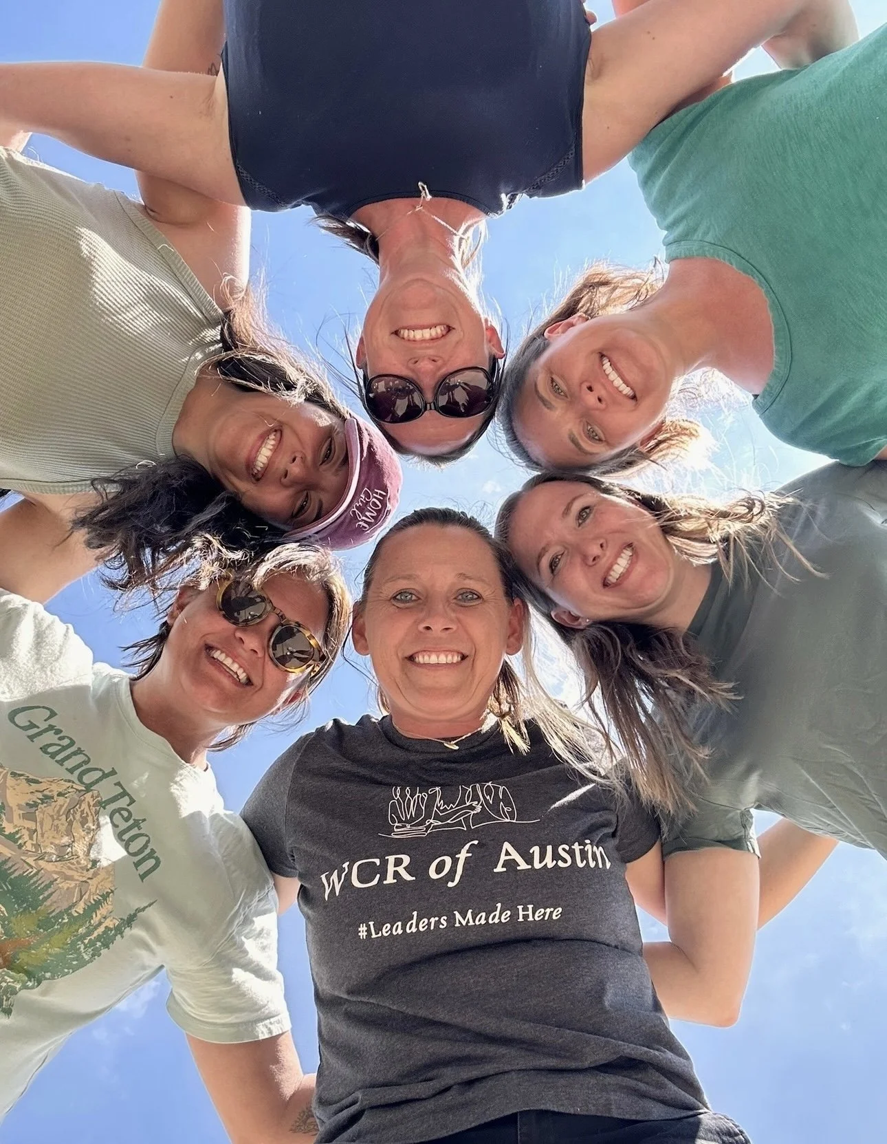 Group of six women looking down into camera, smiling, outdoors on sunny day.