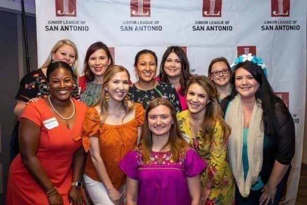 Group of women and girls posing together at a Junior League of San Antonio event, standing in front of a branded backdrop.
