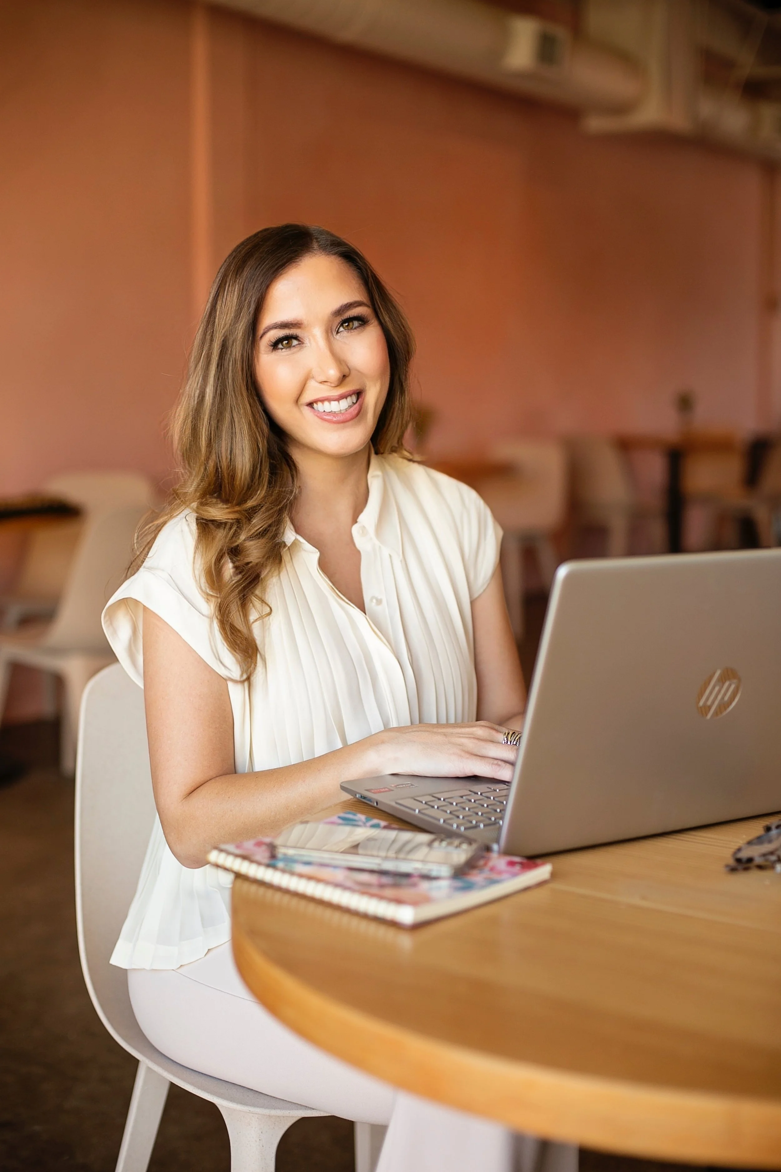 April Guerra with long brown hair, wearing a white pleated blouse, sitting at a round wooden table with a silver HP laptop, notebooks, and a phone, smiling at the camera inside a cozy cafe or coffee shop.
