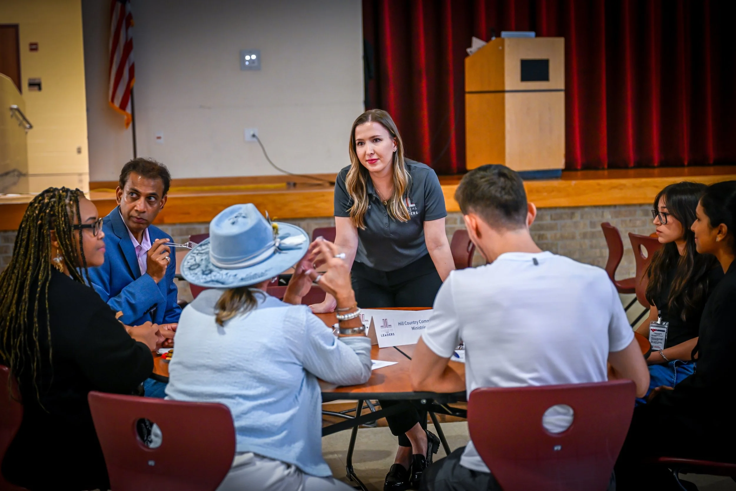 A diverse group of people sitting around a table in a meeting room with a woman standing and talking to them.