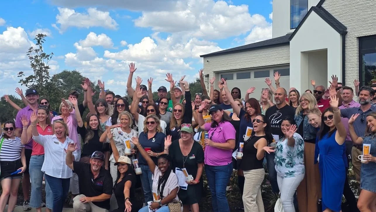 Group of people gathered outdoors, smiling, waving, and celebrating in front of a modern house with a blue sky and clouds above.