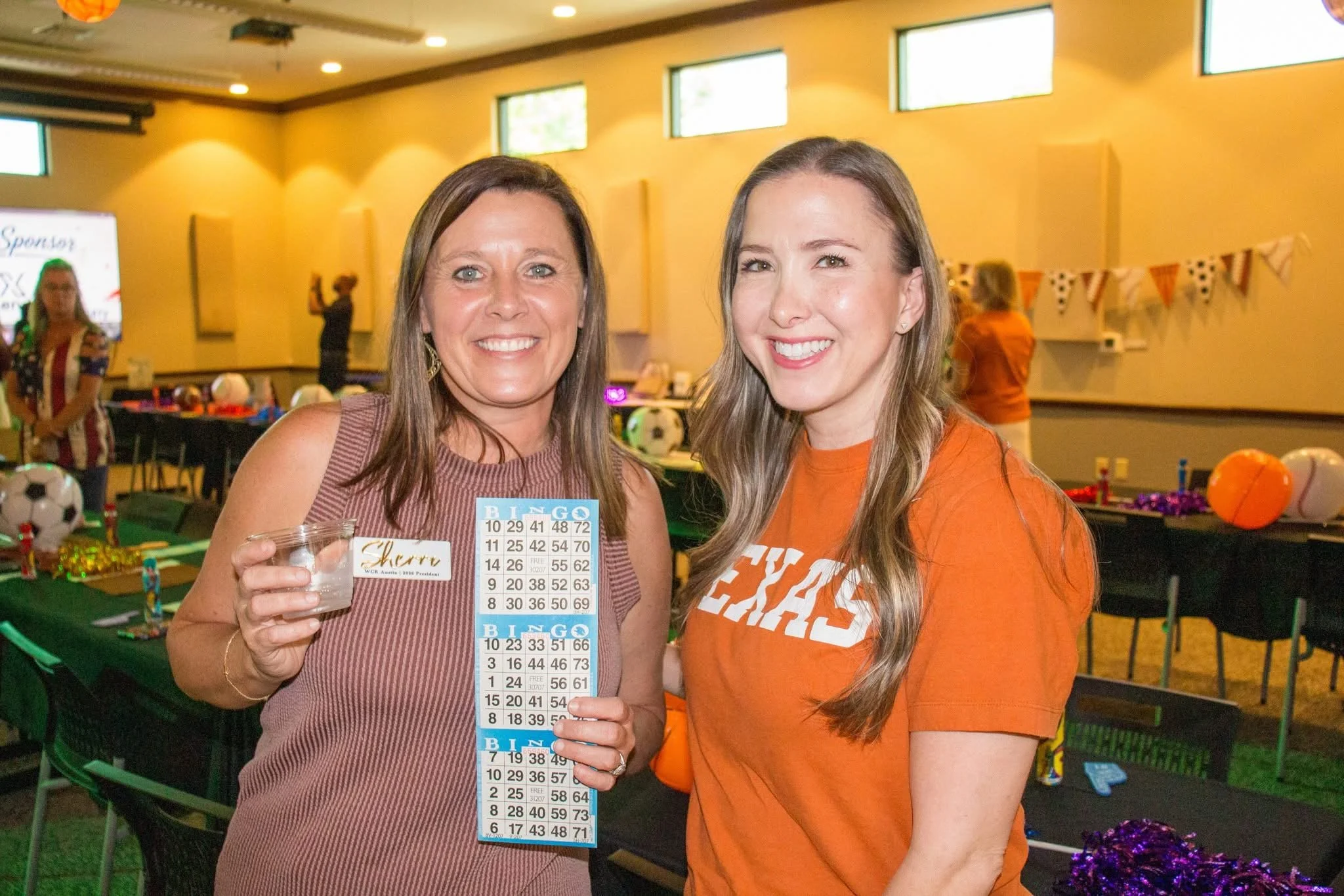 Two women smiling, standing indoors at a bingo event with tables, balloons, and decorations in the background. The woman on the left holds a small cup and a bingo card, the woman on the right wears an orange shirt.