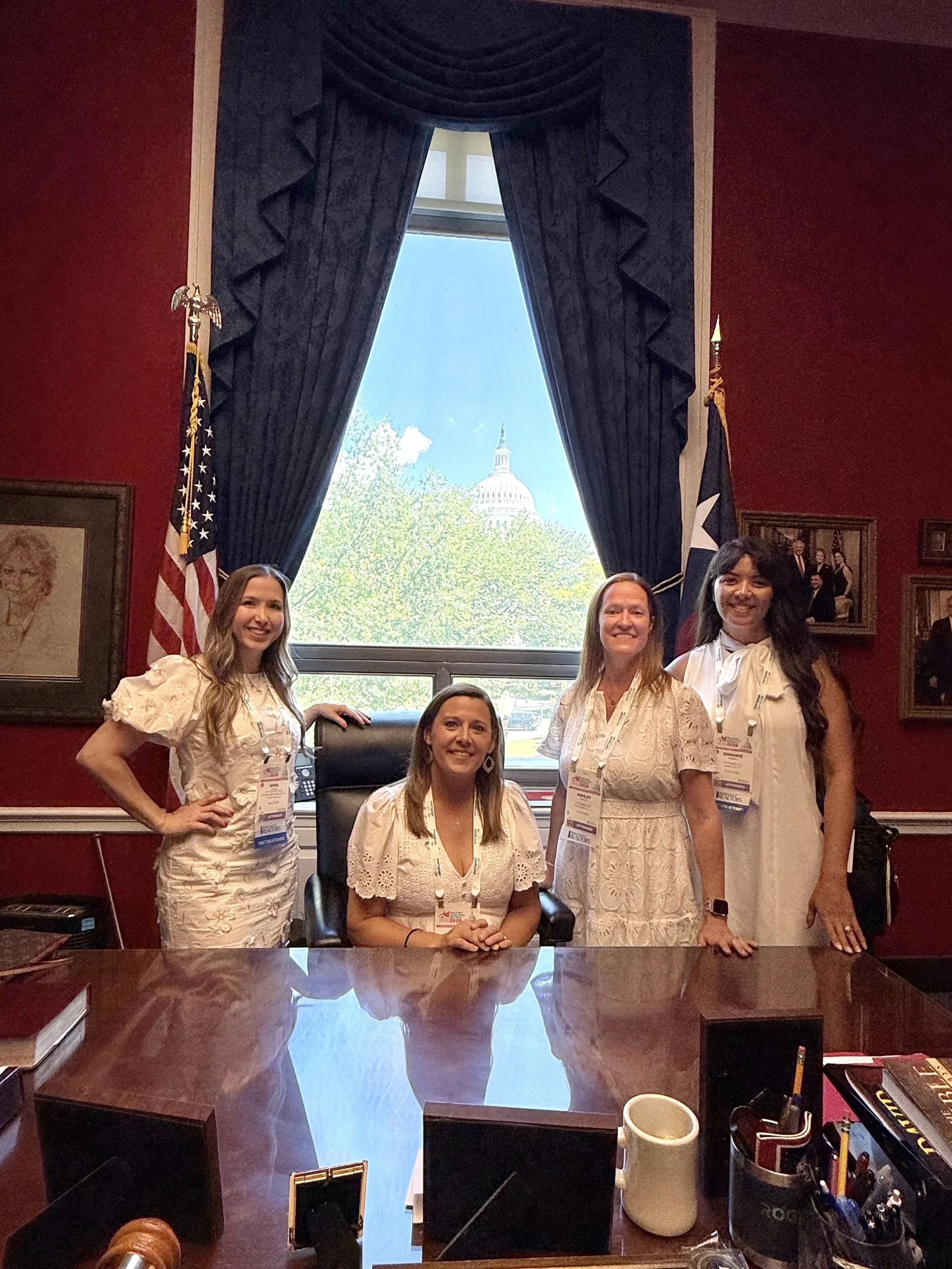 Four women in business attire posing in a conference room with a large window showing the U.S. Capitol building and green trees outside. The room has dark red walls, framed pictures, and American flags. There are office supplies and a coffee mug on t