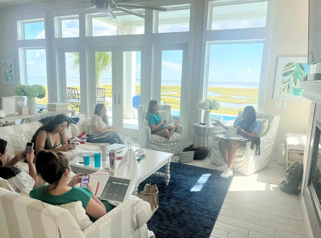 Group of women sitting in a bright living room with large windows and a view of marshland and water outside. Some women are using their phones, one is taking notes, and two are engaged in conversation. The room is decorated with white furniture, flow