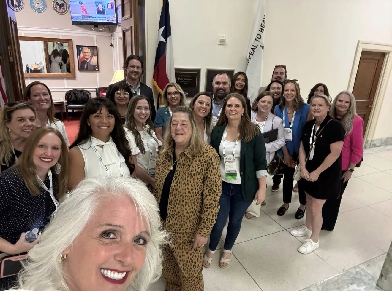 Group of diverse people smiling for a selfie in a room with flags and a TV screen in the background.