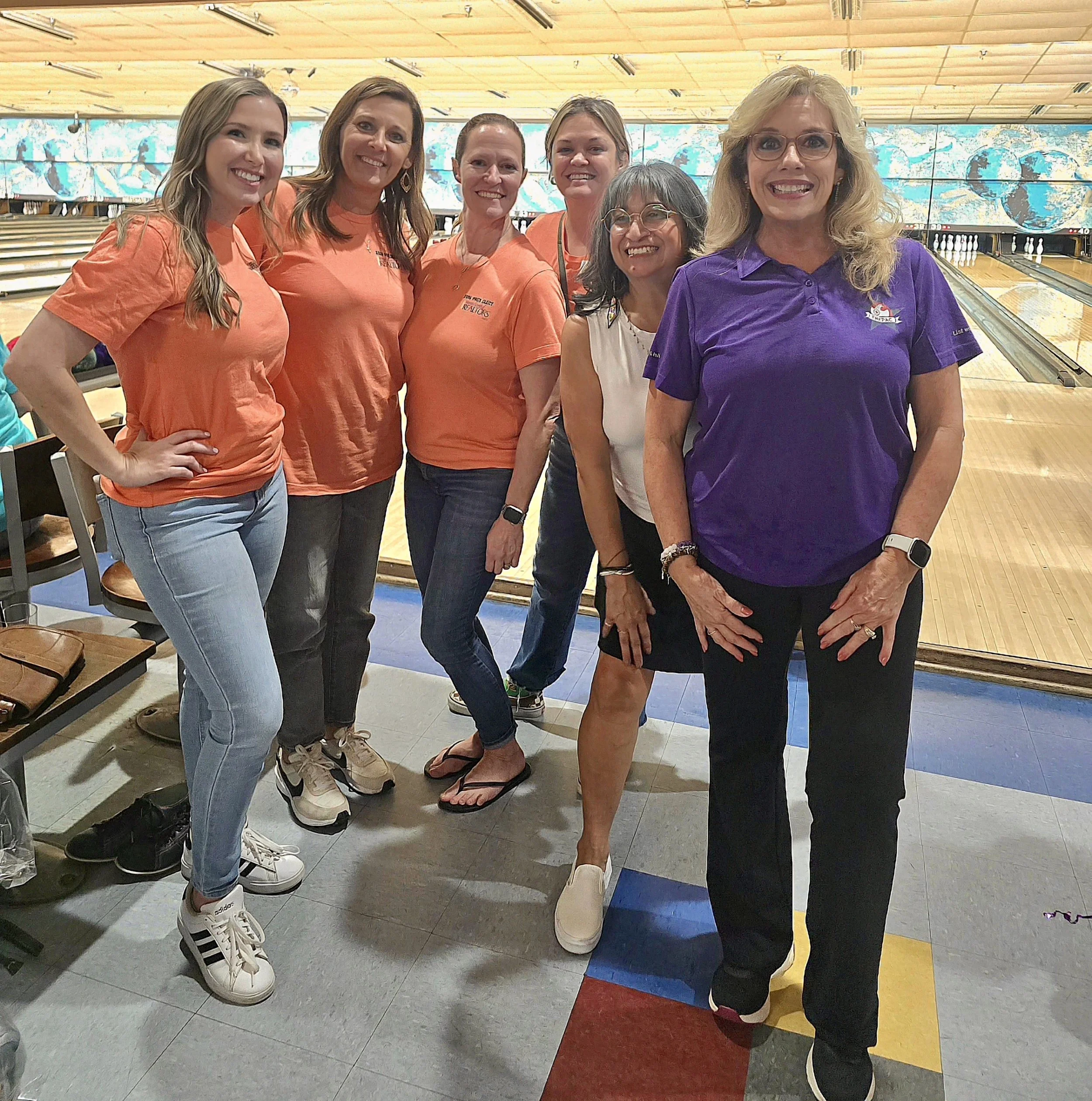 Group of women standing and smiling at bowling alley, some wearing matching orange shirts, one woman in purple shirt, with bowling lanes and large screen in the background.