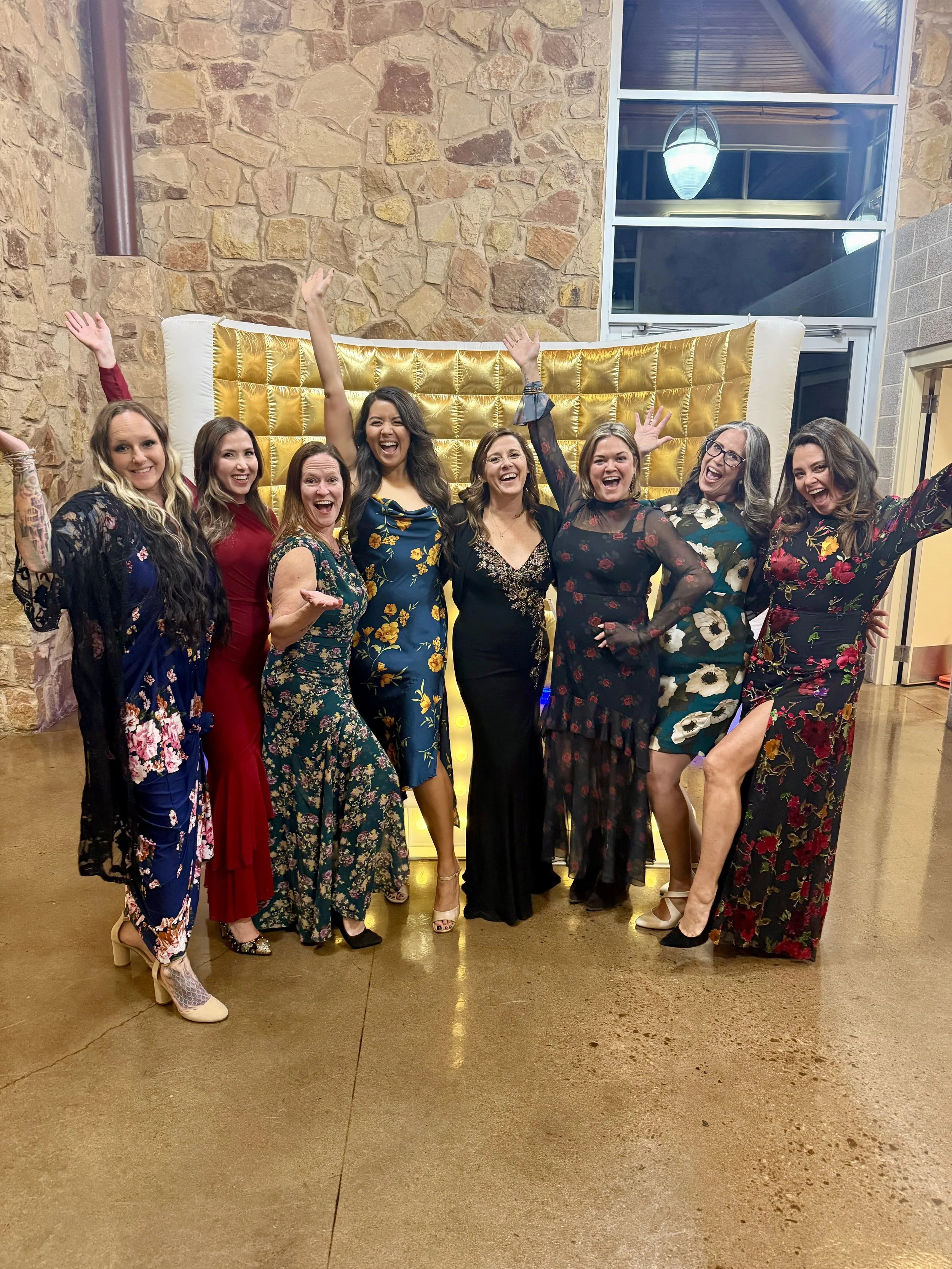 A group of nine women dressed in colorful dresses, smiling and celebrating indoors against a stone wall and large window, with a yellow backdrop behind them.