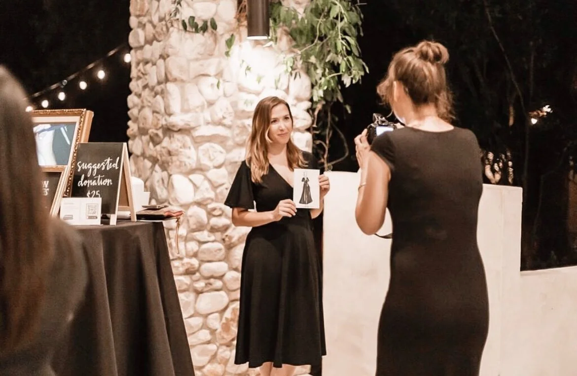 April Guerra in black dress posing for a photo at a nighttime event, holding an illustration of a gown, while another woman takes her picture, surrounded by decorative string lights and a stone wall.