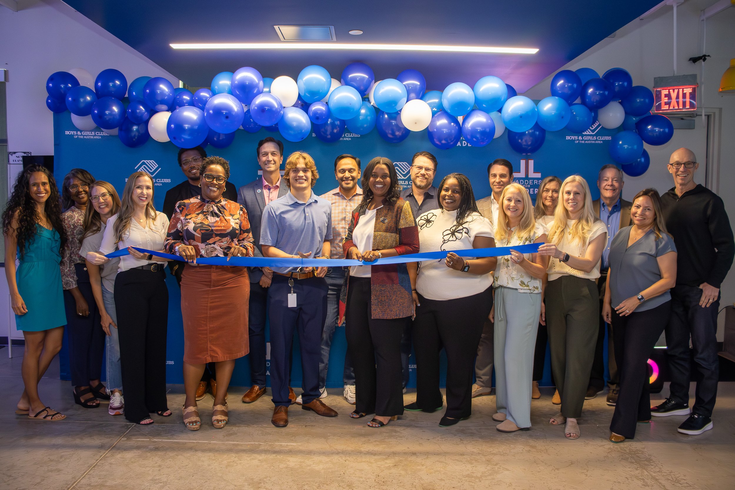 A diverse group of people posing together at an event, with some holding a blue ribbon for a ribbon-cutting ceremony. They are standing in front of a blue backdrop decorated with blue and white balloons, and the backdrop has the logo of Boys & Girls 
