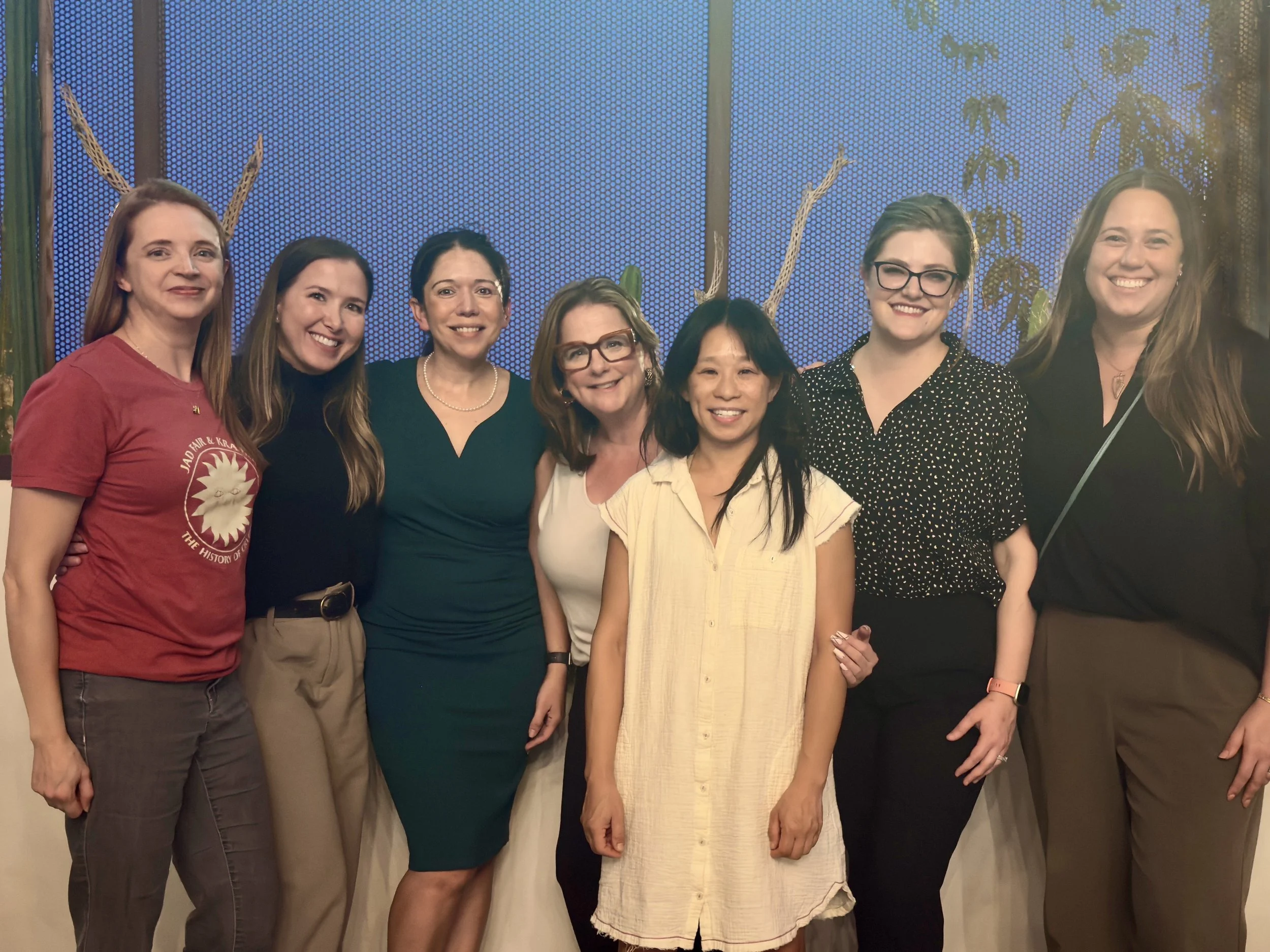 Group of seven women standing together indoors, smiling for the camera, with a blue patterned background and some plant leaves visible behind them.