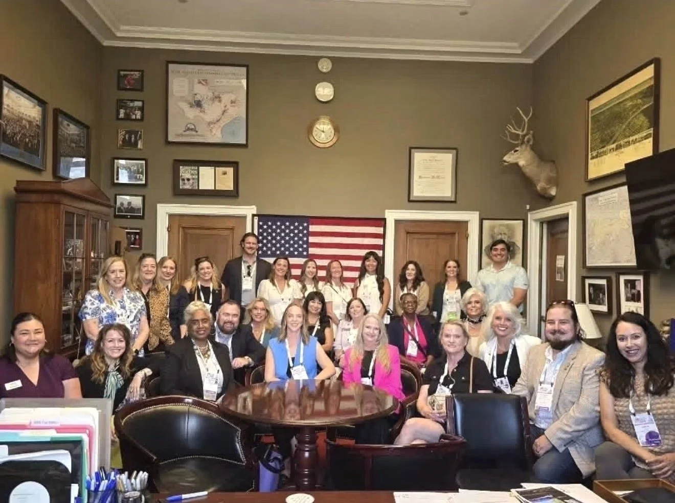 Group of diverse people gathered in a room with American flag, framed pictures, and a mounted deer head on the wall, posing for a photo.
