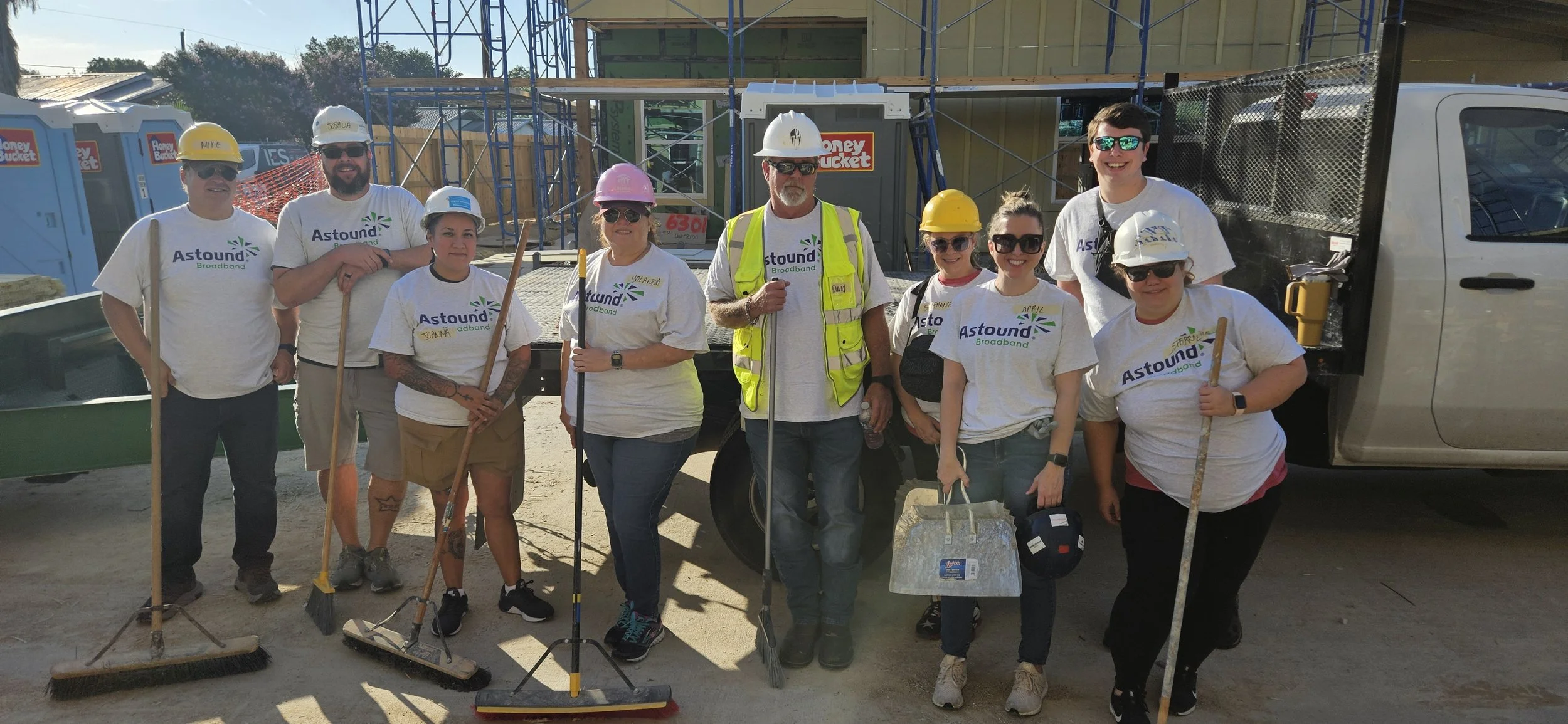 Group of ten people at a construction site wearing hard hats, safety vests, and white T-shirts with the Astound Broadband logo, standing in front of construction equipment and a partly constructed building.