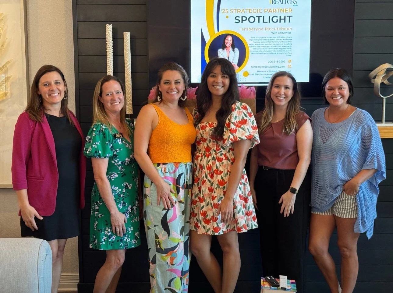 A group of six women standing together indoors, smiling at the camera in front of a large screen displaying a presentation about a strategic partner spotlight, featuring a woman and contact information.