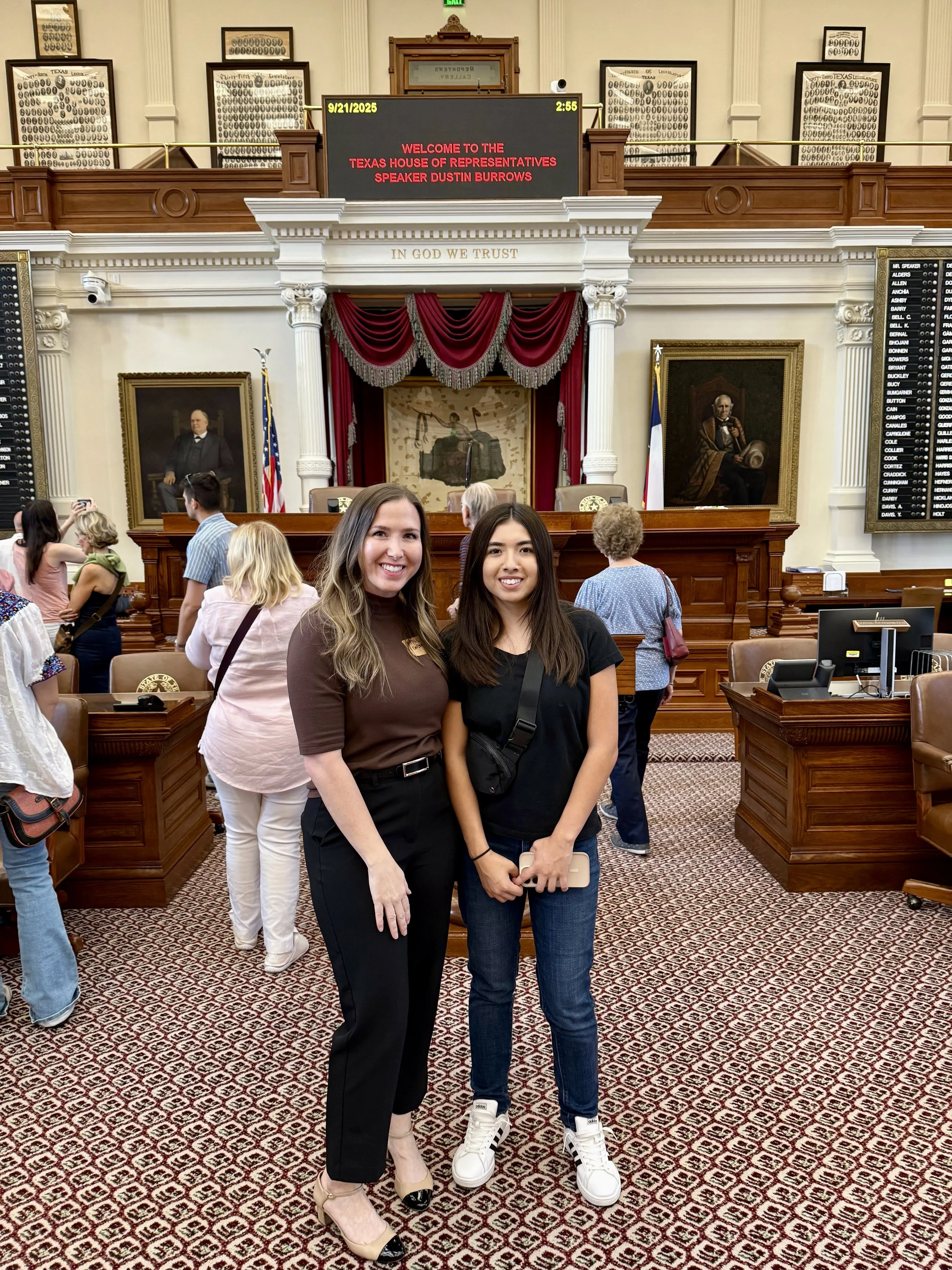 Two women standing inside the Texas House of Representatives chamber, smiling at the camera. Other people are in the background, some taking photos. The chamber features portraits of historical figures, a digital display board with the date, time, an