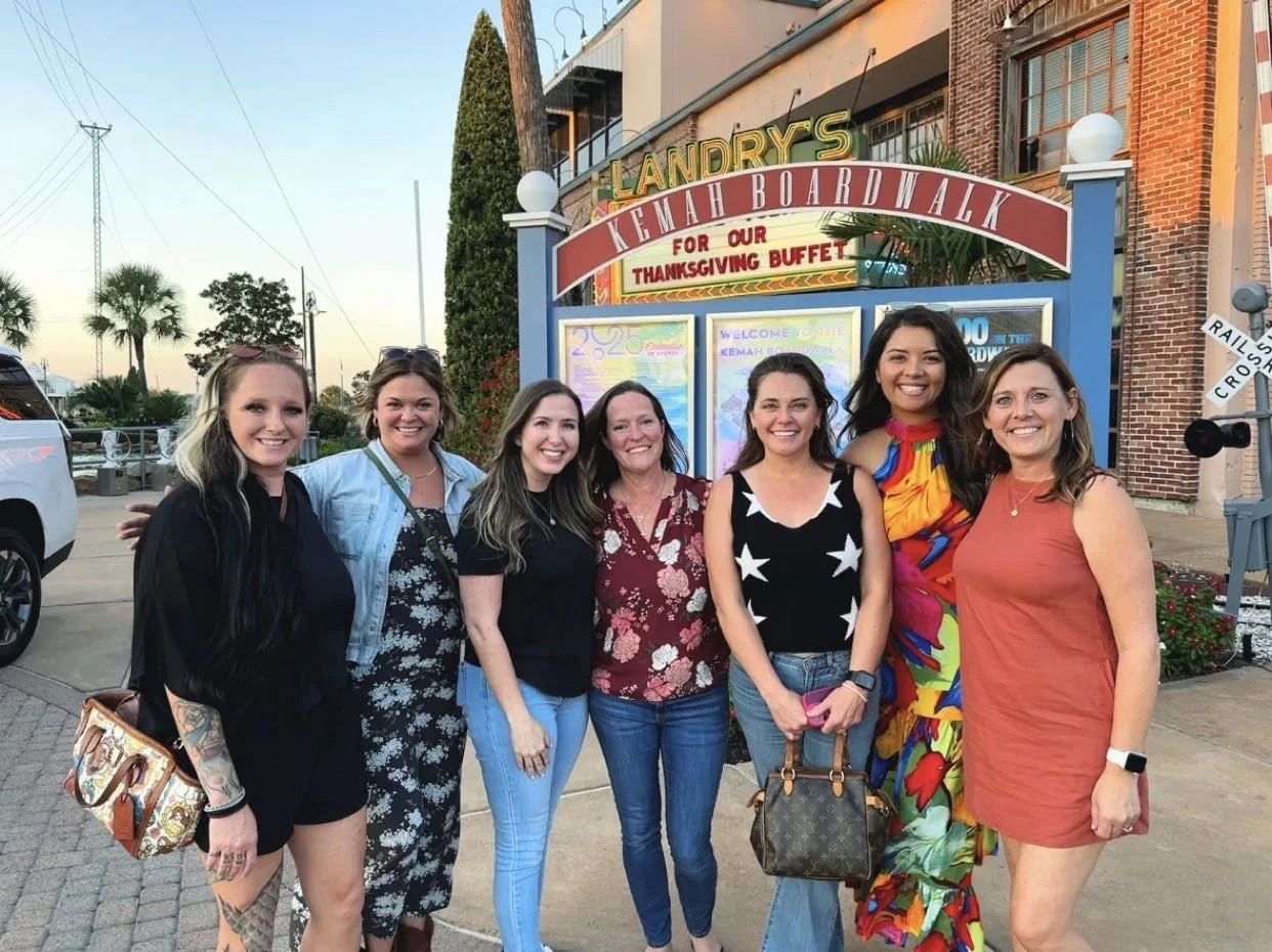 Group of seven women standing in front of a sign for Landry's Kemah Boardwalk, smiling at the camera.