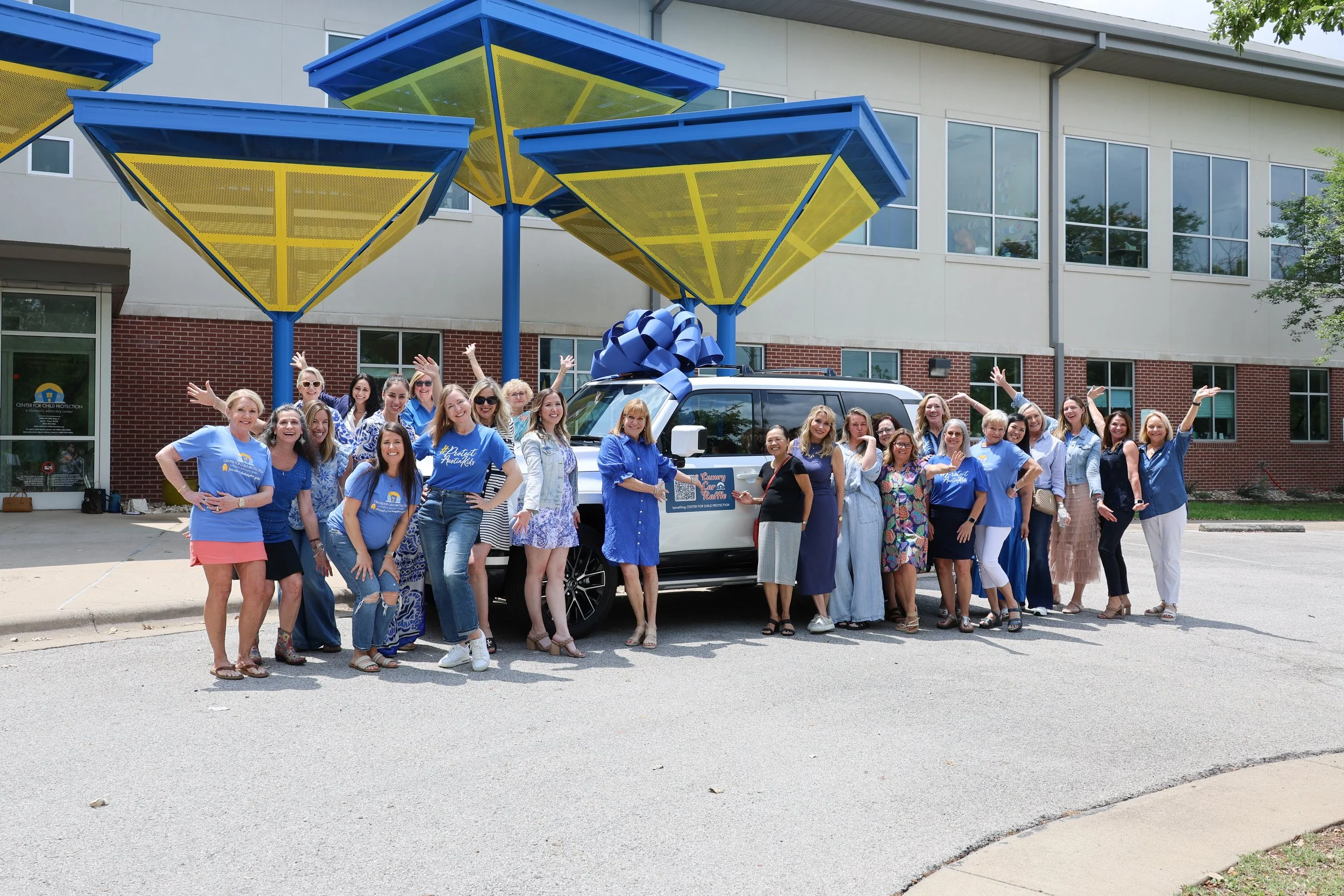 Group of women celebrating in front of a large wrapped vehicle with a big blue ribbon. The women are dressed in blue and summer clothing, standing outdoors in front of a modern building with colorful structures above. Everyone is smiling and raising