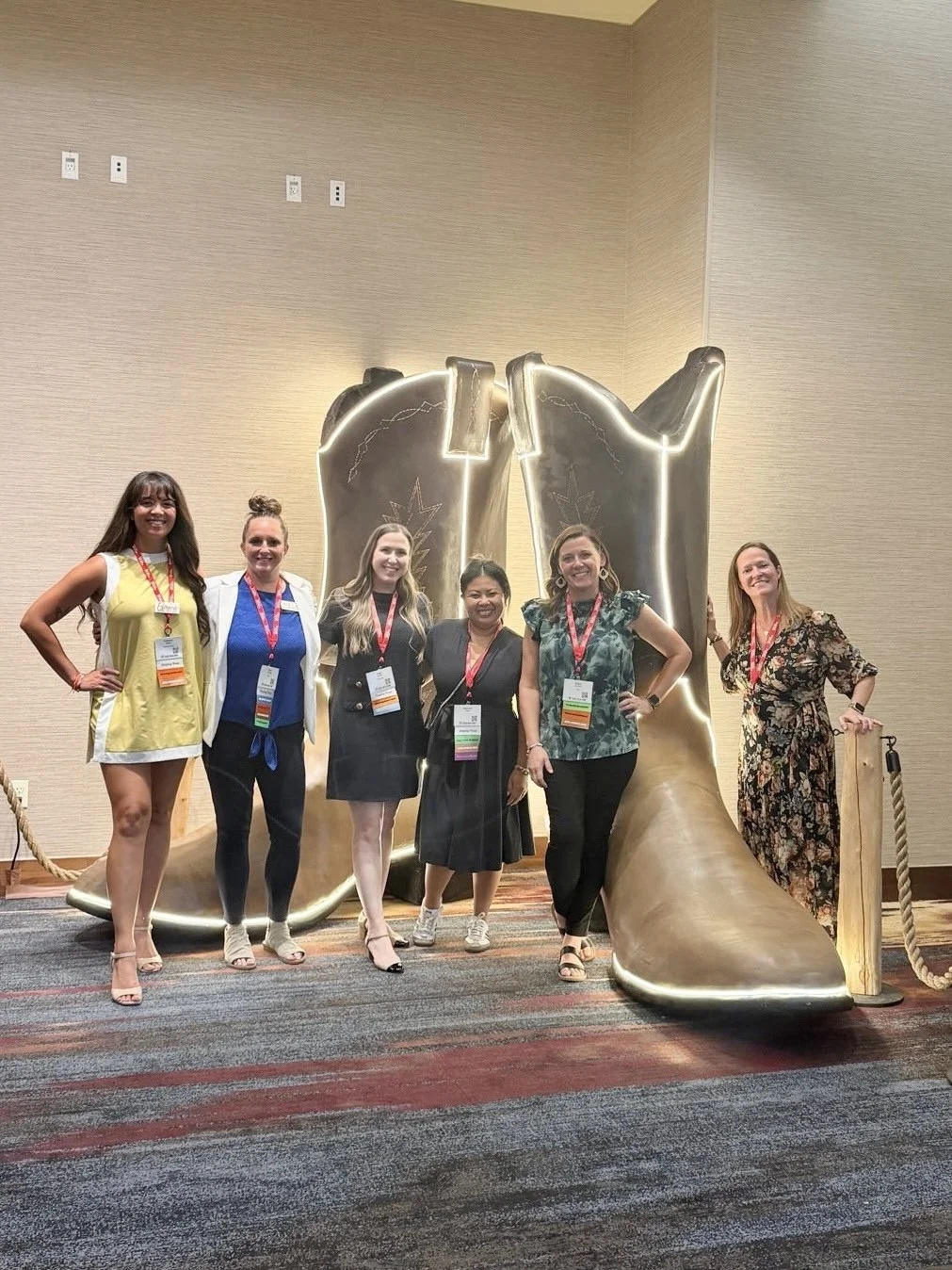 Group of six women standing in front of a large, metallic cowboy boot sculpture at a conference.
