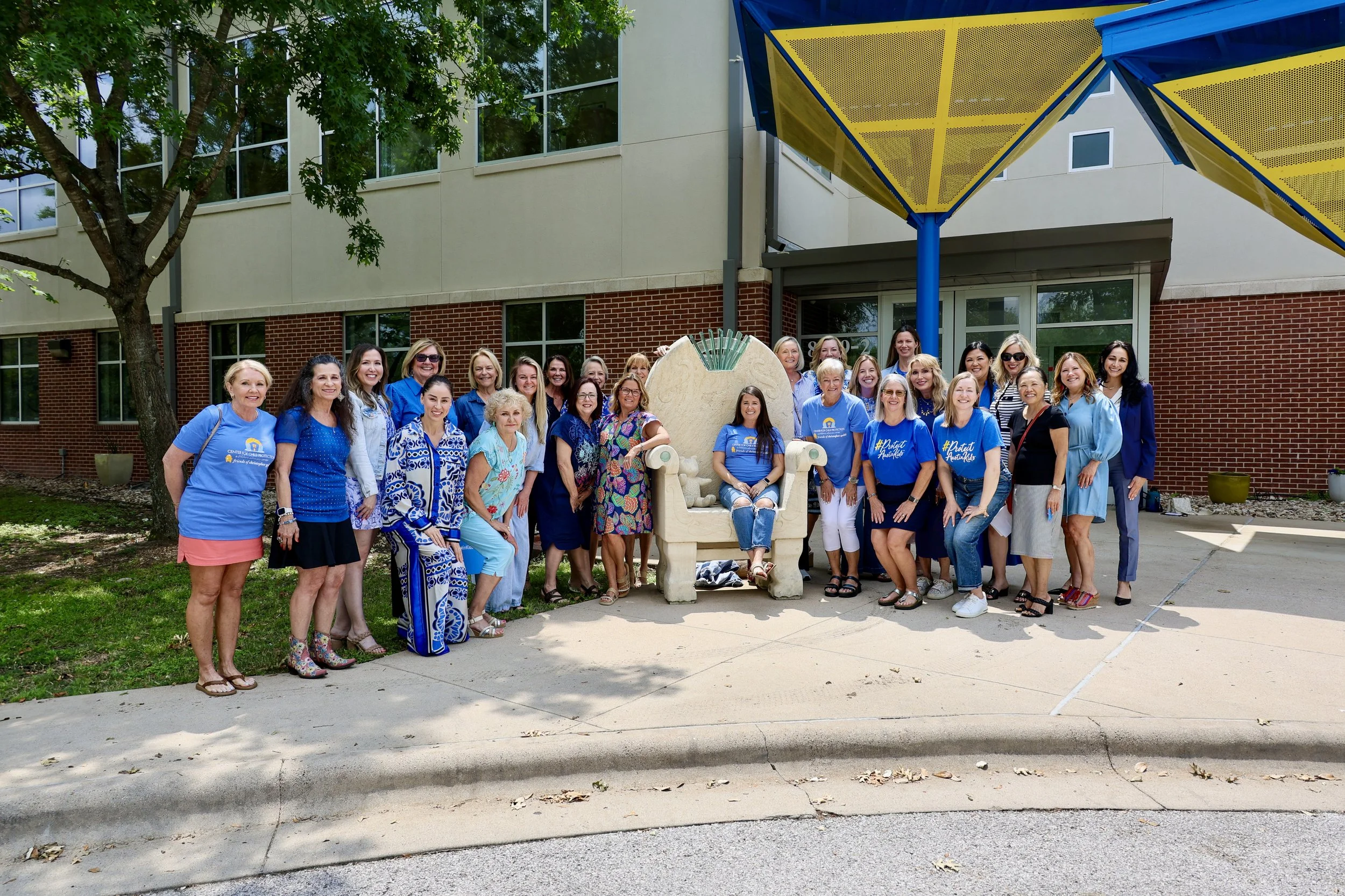 Group of women standing outdoors in front of building, posing around a large decorated chair with a woman seated in it, some women wearing blue shirts with design, trees and colorful canopy overhead.