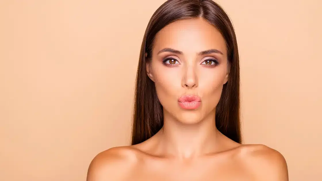 Close-up of a woman with long brown hair, making a kissy face pose, against a beige background.