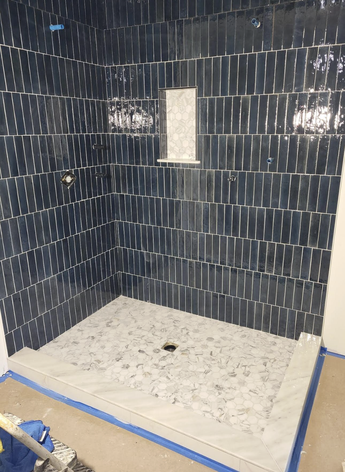 A shower with dark blue tile walls, a small built-in shelf, and a marble tiled floor with a drain in the center.