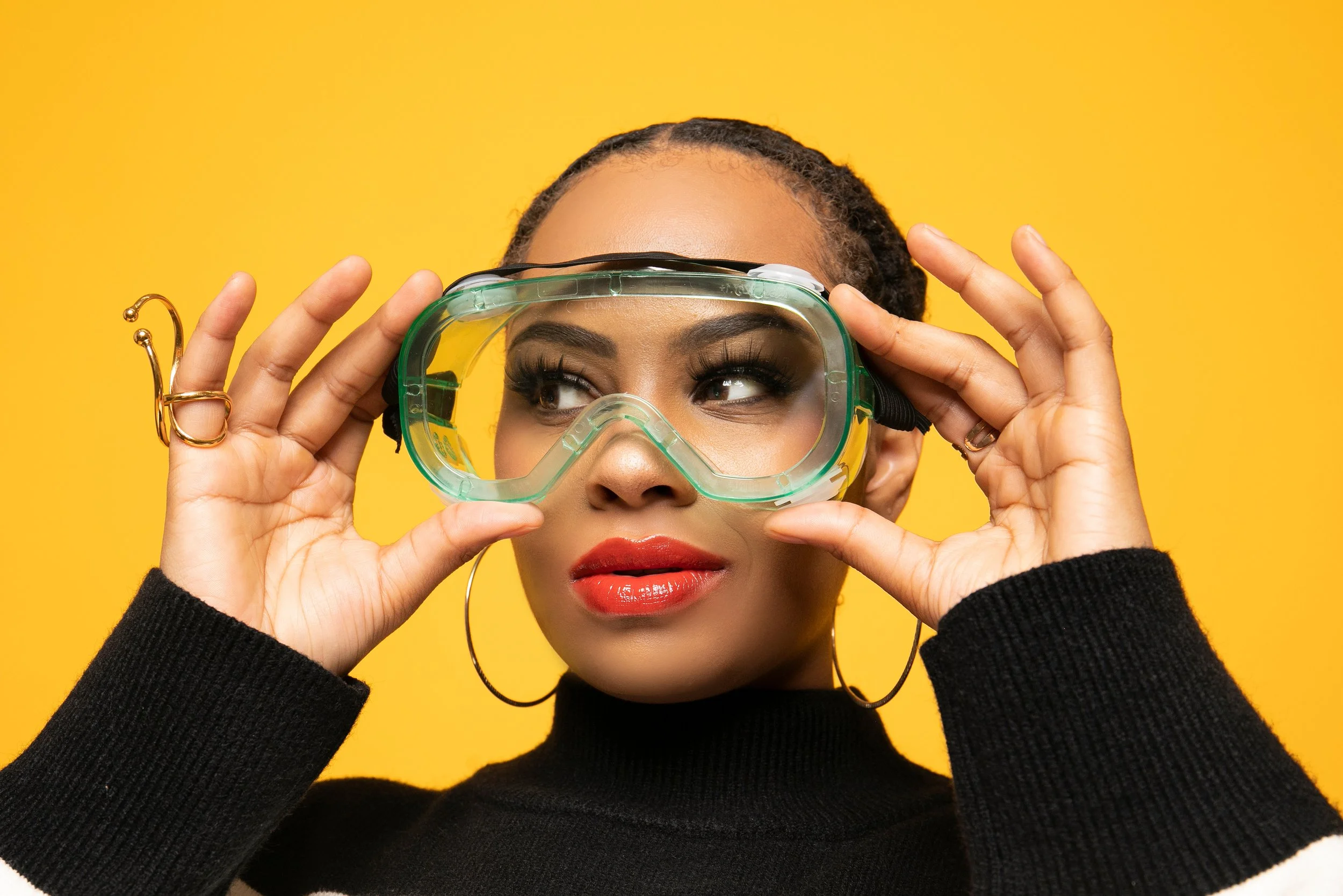A woman with dark skin, wearing a black long-sleeve top and gold hoop earrings, adjusting yellow swimming goggles on her face against a bright yellow background.