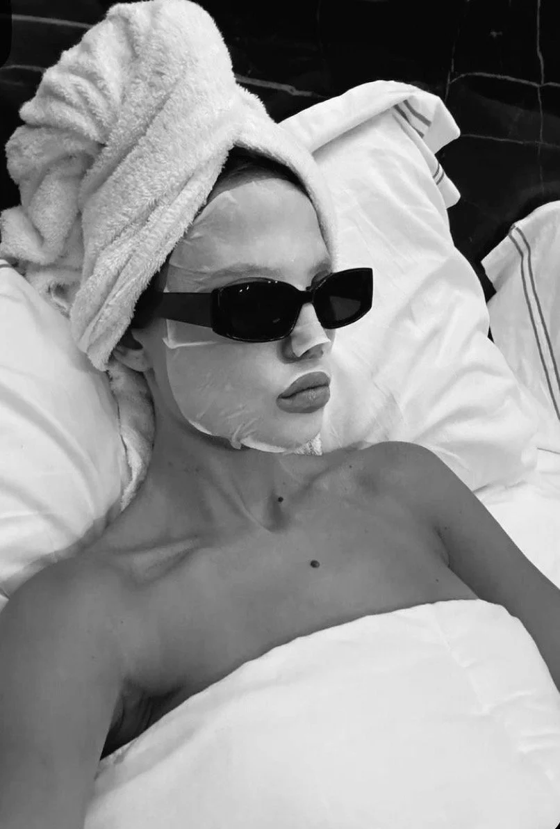 A person lying in bed with a towel wrapped around their head, wearing large sunglasses, a facial mask, and lipstick, with a pillow and blanket partially visible.