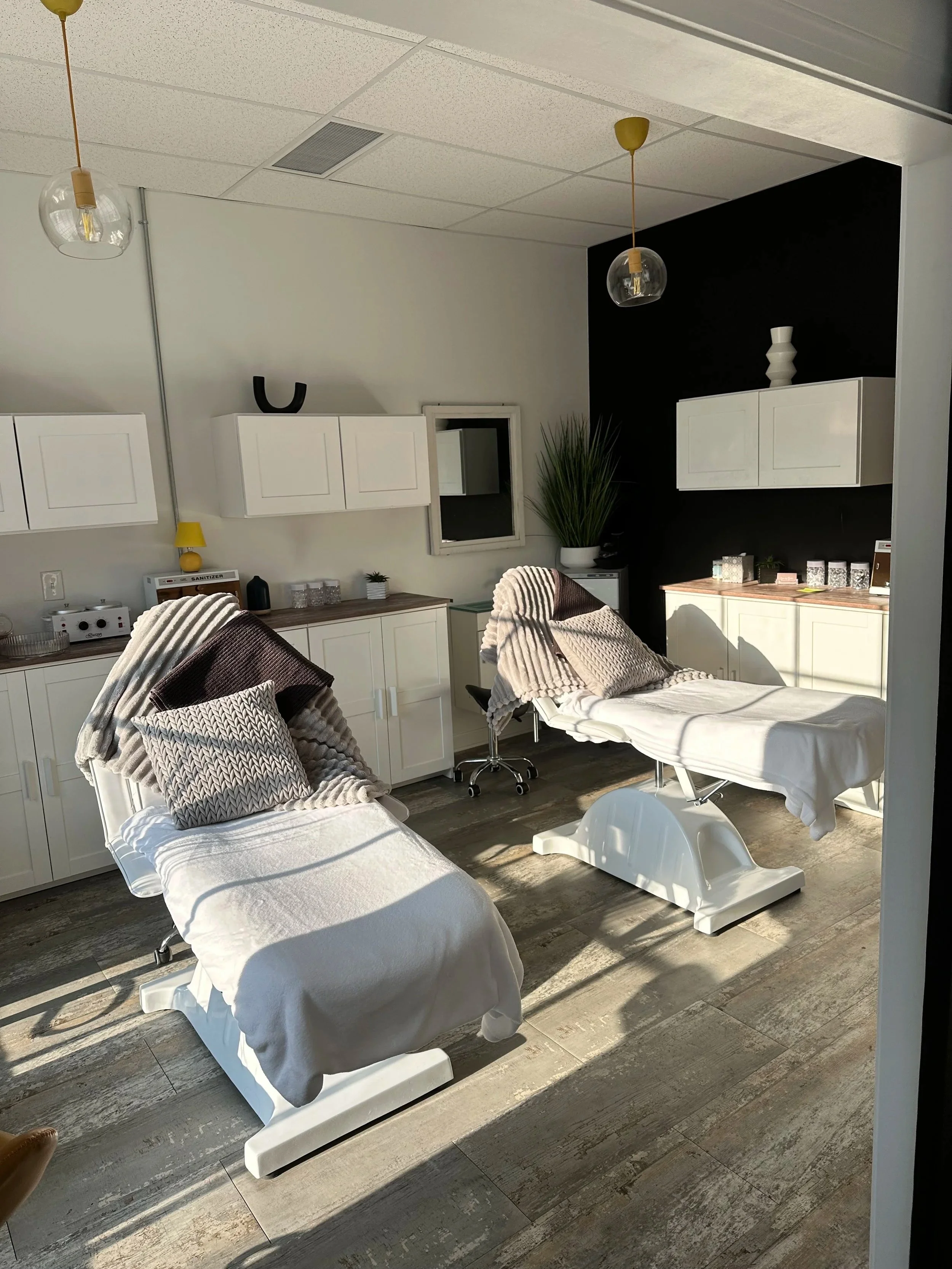 Two treatment tables covered with blankets and pillows in a modern health clinic or massage room, with cabinets, decorative items, and a tall plant in the background.