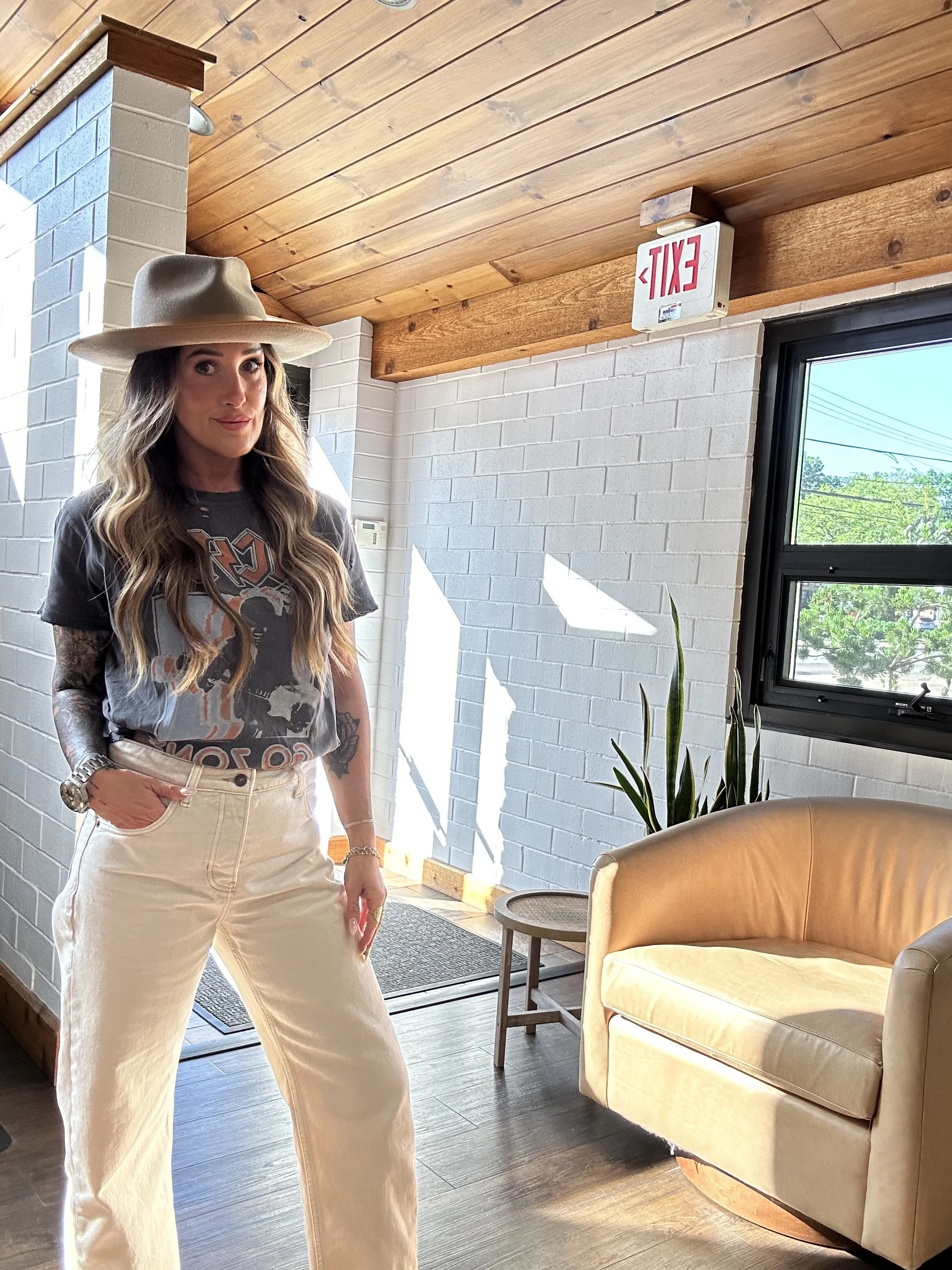 Woman with long wavy hair wearing a wide-brimmed hat, graphic T-shirt, and cream-colored pants standing in a brightly lit room with white brick walls, a window, a beige armchair, and a small round side table.