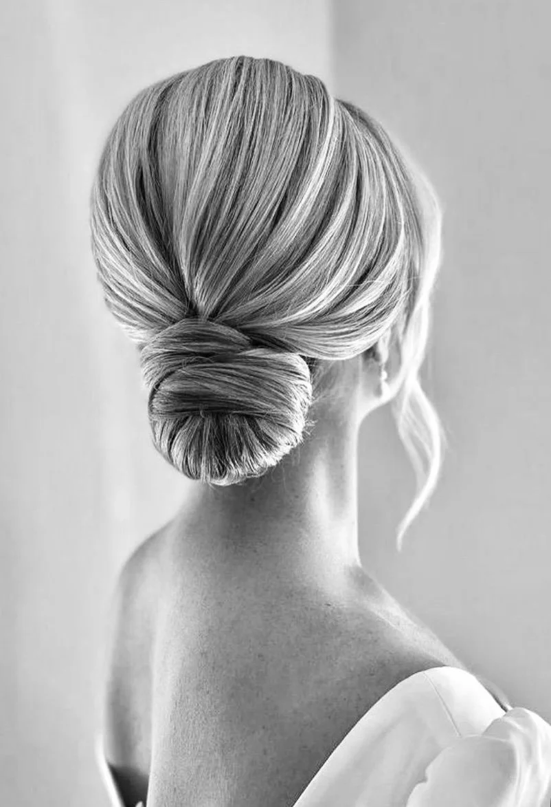 A black and white photograph of a woman with blonde hair styled in a sleek, elegant bun at the nape of her neck, with a loose strand hanging down.
