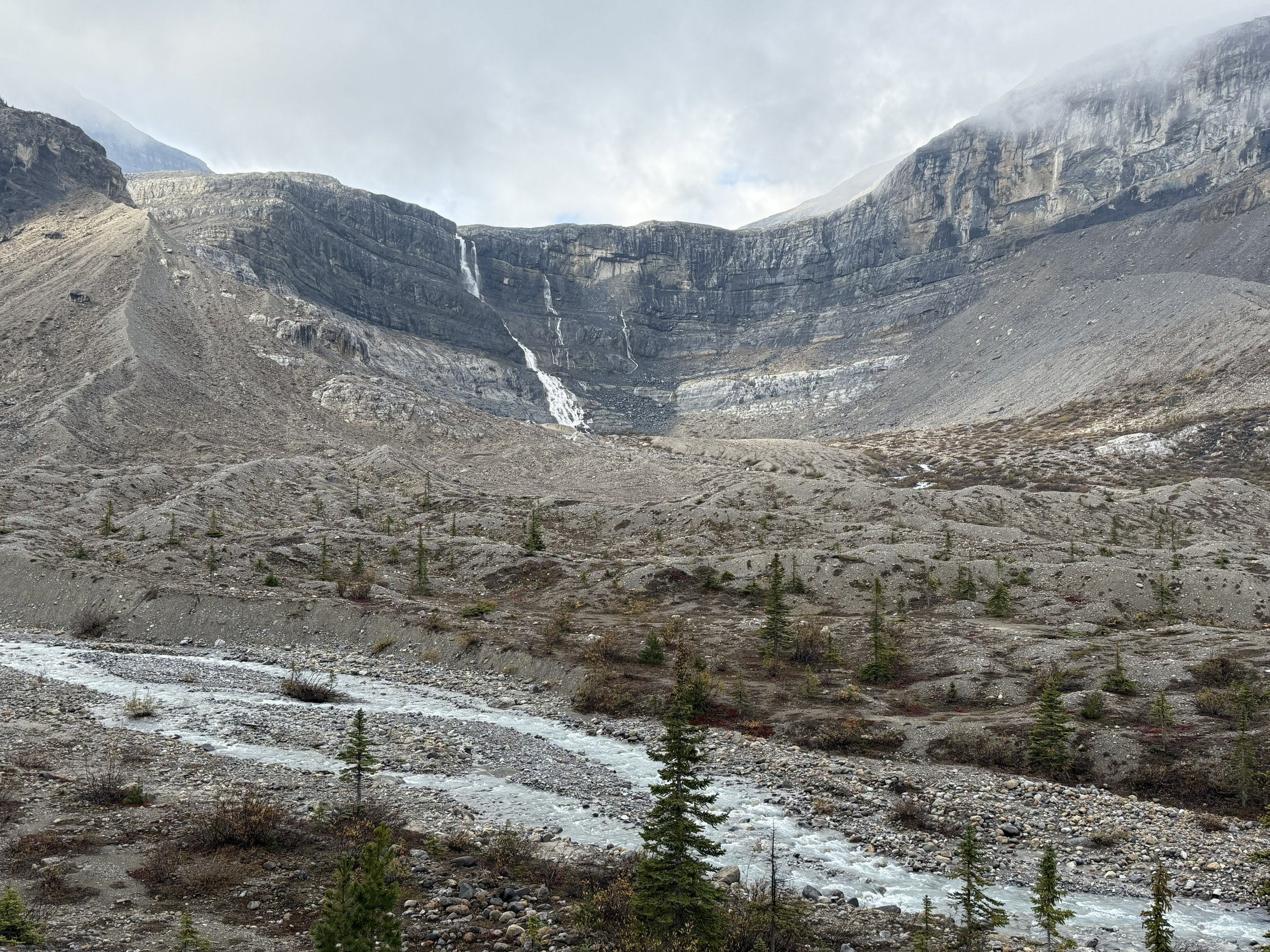 Mountain landscape with steep rocky slopes, a small waterfall, evergreen trees, and a flowing river at the bottom.