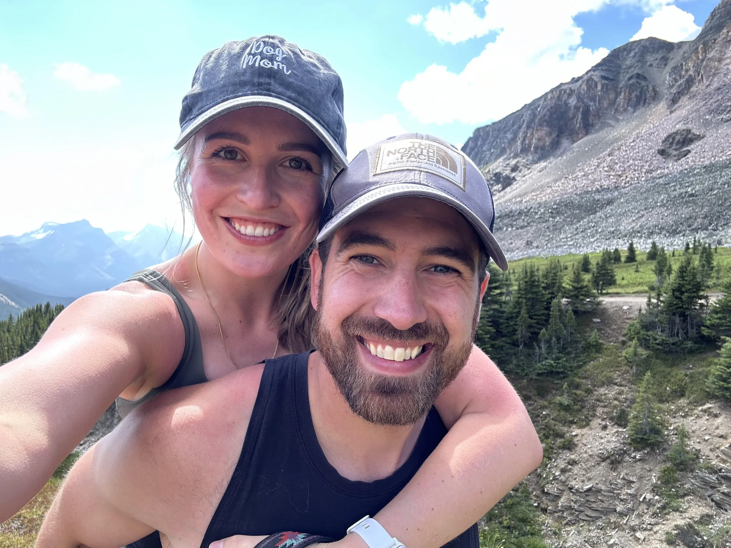 A smiling couple taking a selfie outdoors with mountain scenery and trees in the background, wearing caps and athletic clothing.