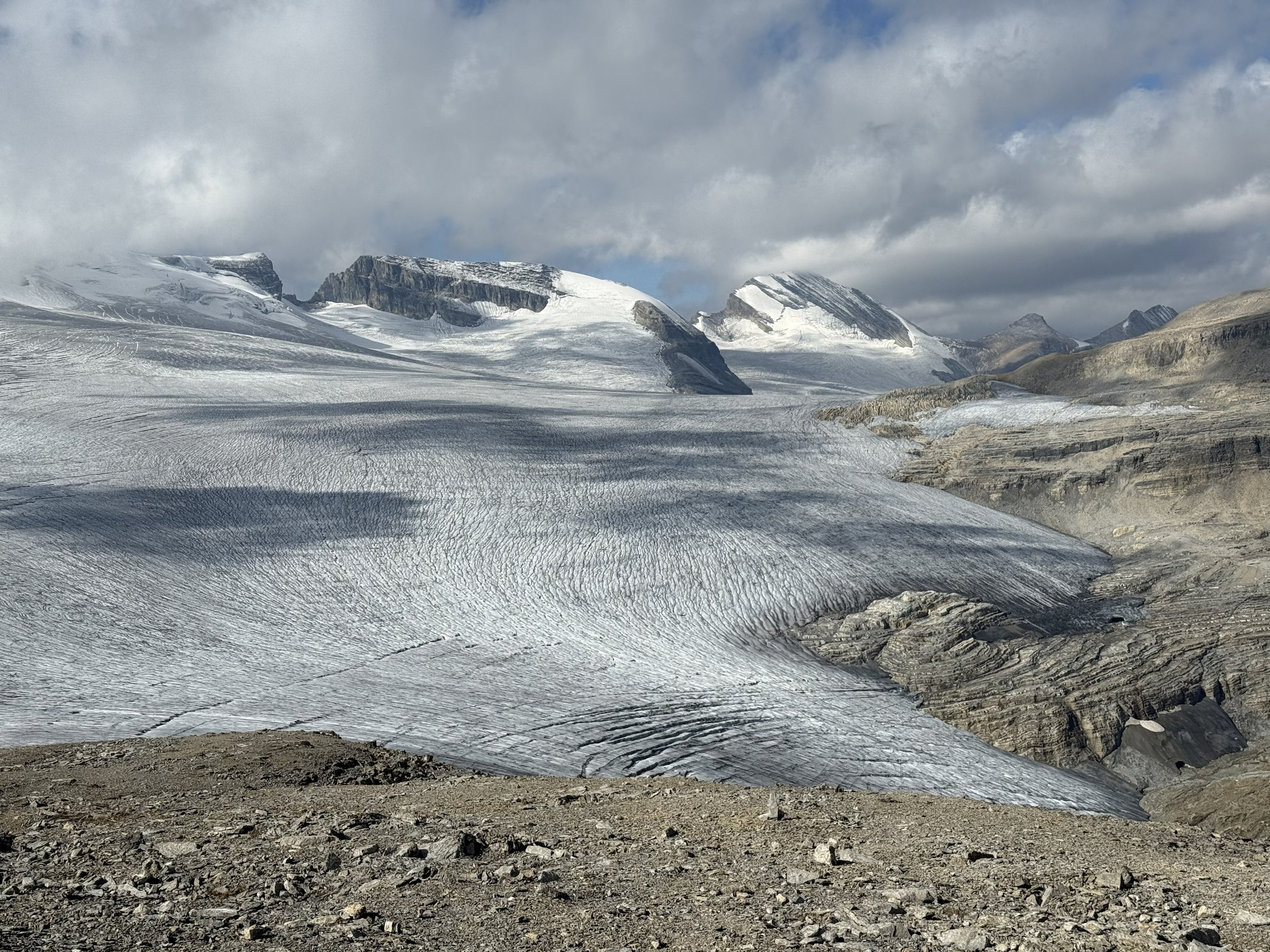 A glacier in a mountainous landscape with snow-capped peaks, rocky terrain, and cloudy sky.