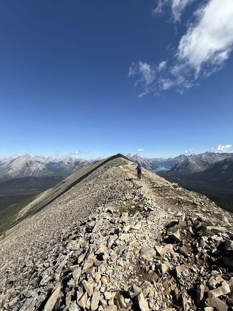 Hiker walking along a rocky mountain ridge under a blue sky with some clouds, with mountain ranges and a river in the distance.