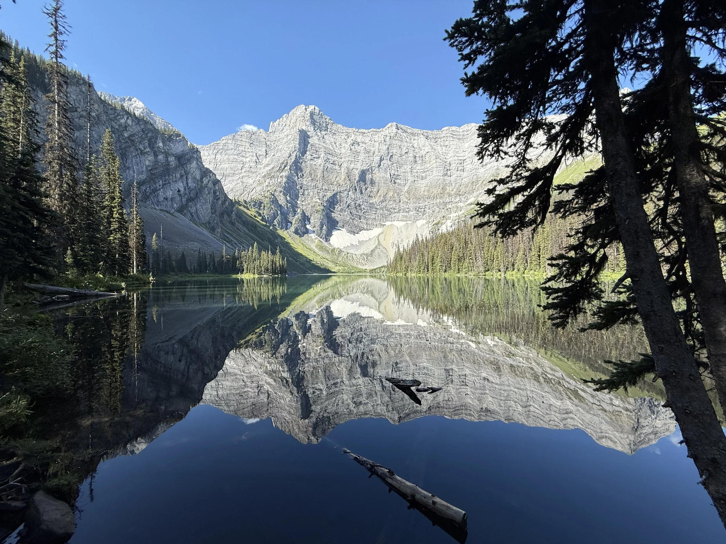 A calm mountain lake reflecting tall, jagged mountains and pine trees under a blue sky.