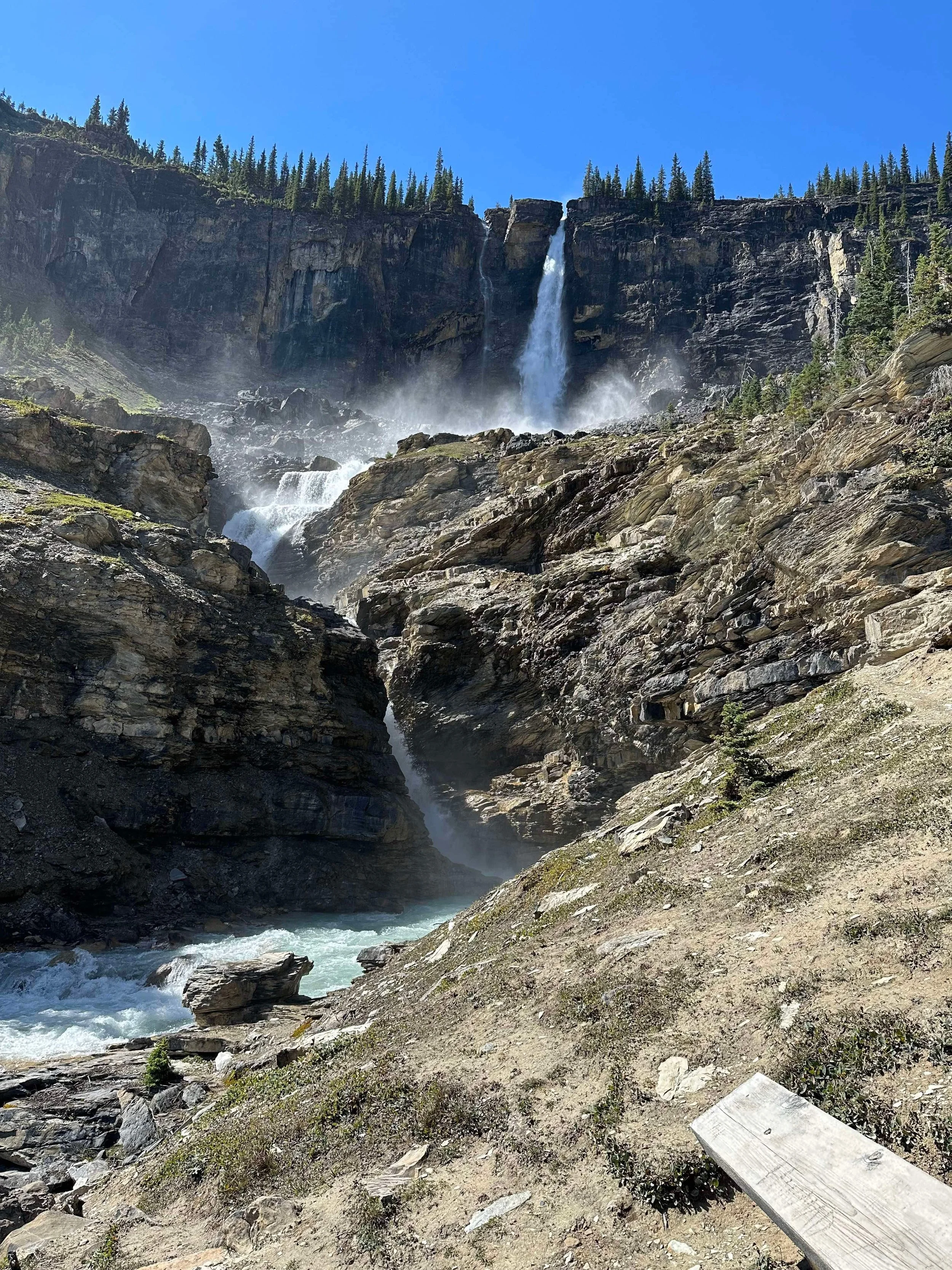 A waterfall flowing down a rocky mountain terrain into a river at the bottom, with trees and a clear blue sky in the background.