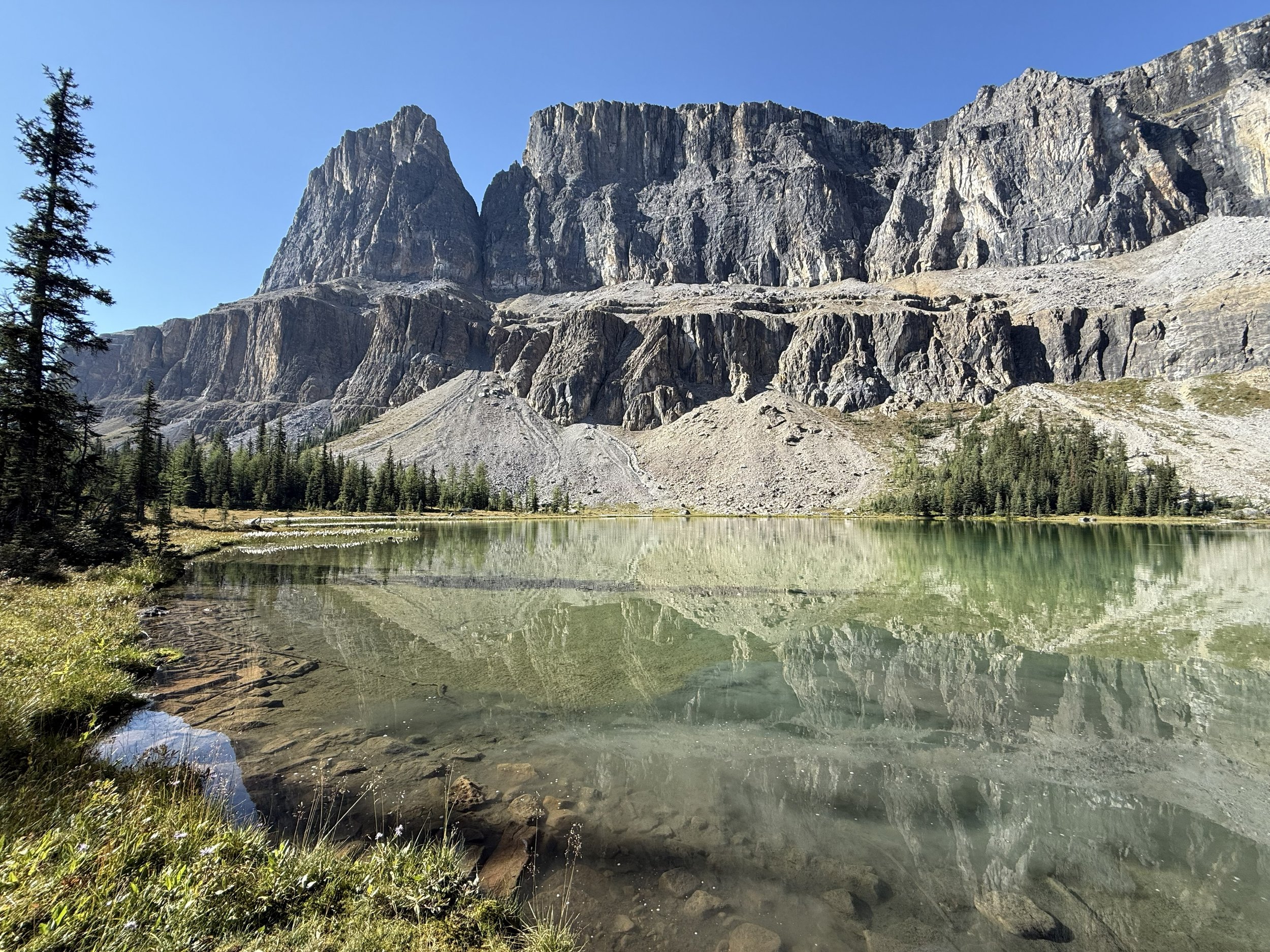 Mountain lake with clear water reflecting tall, rocky mountains and green pine trees under a blue sky.
