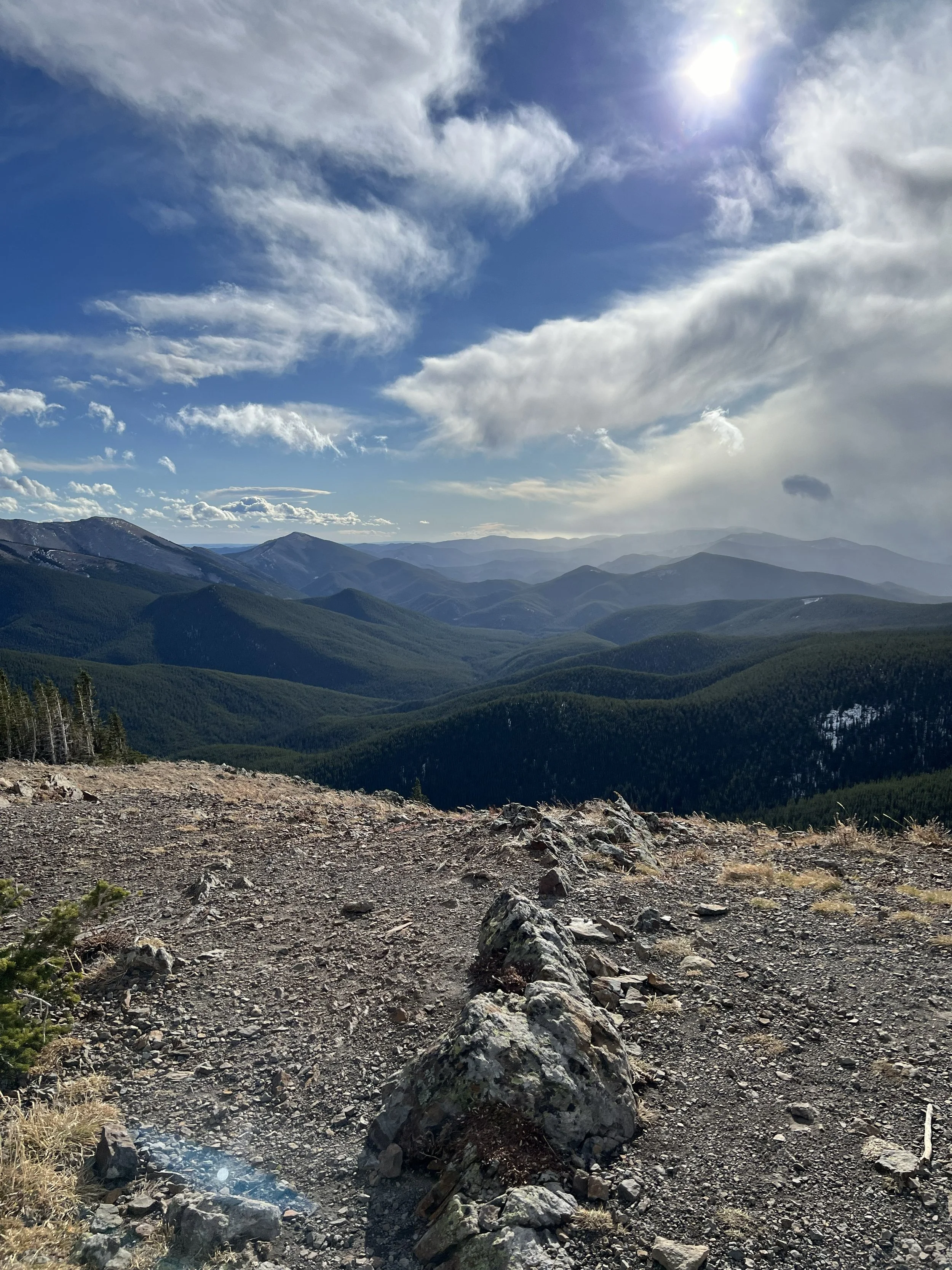 A scenic view of mountain ranges covered with dense green forests under a partly cloudy sky, with the sun shining brightly.