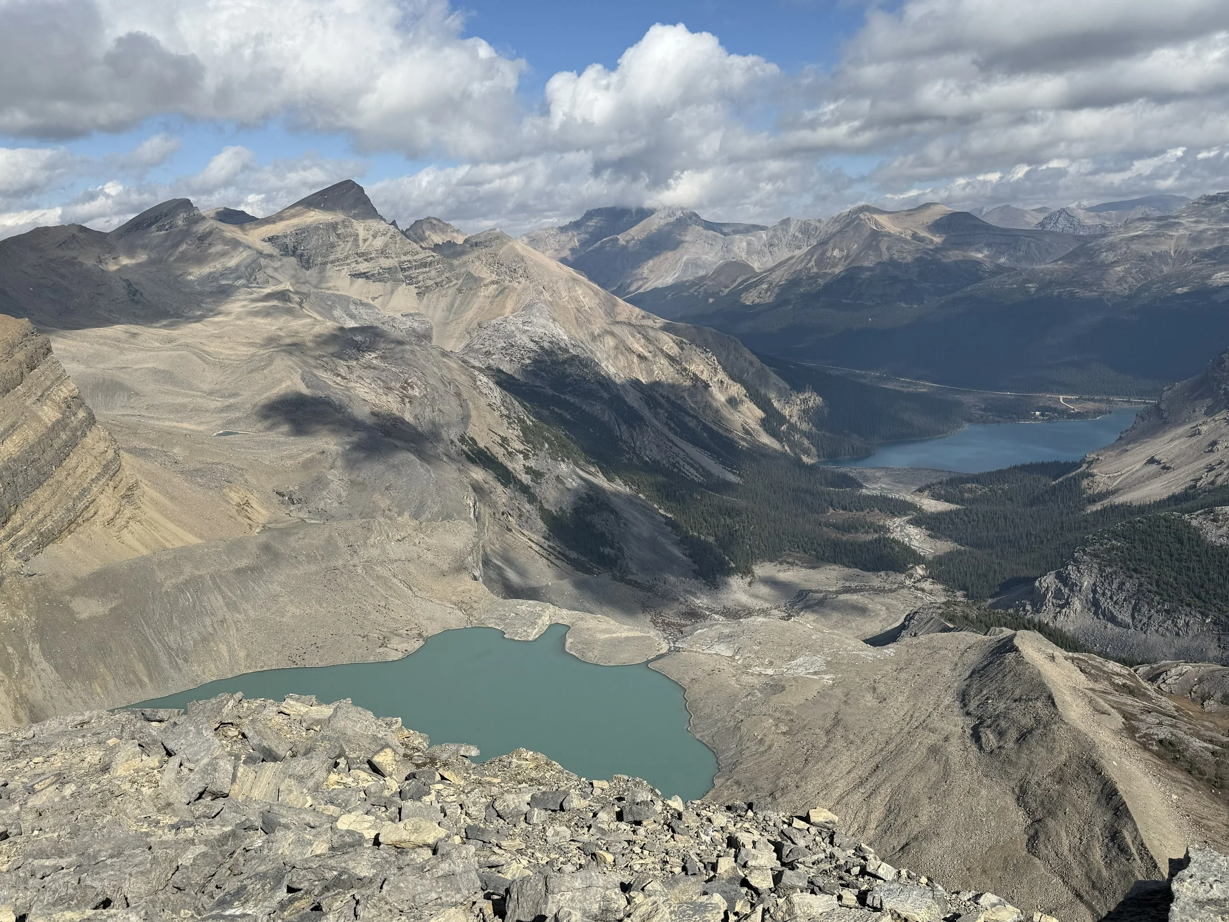 Mountains and rocky terrain with two blue lakes, one in foreground and one further back, under cloudy sky.