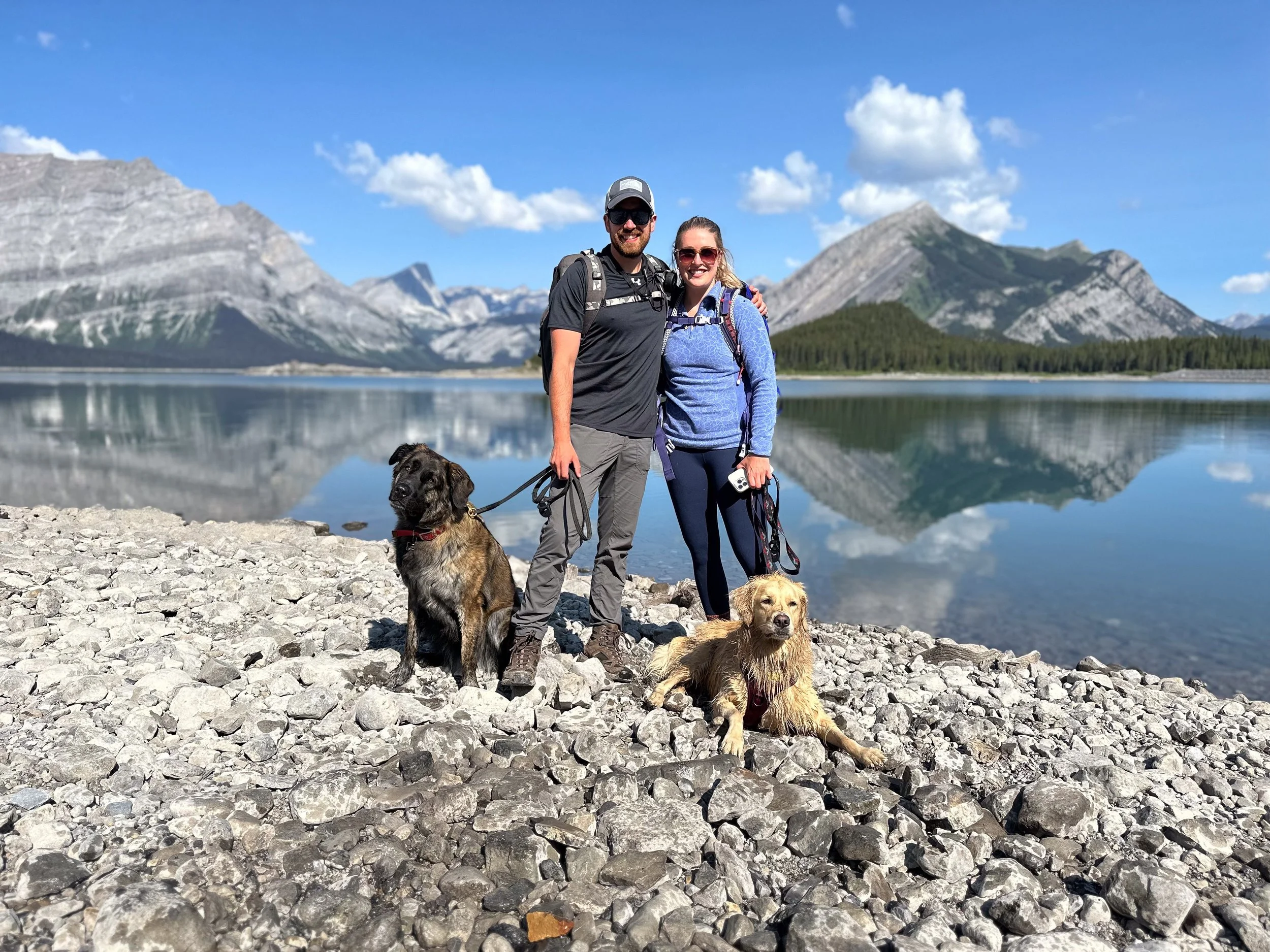A couple with two dogs stands on a rocky shore next to a mountain lake with mountains and a blue sky in the background.