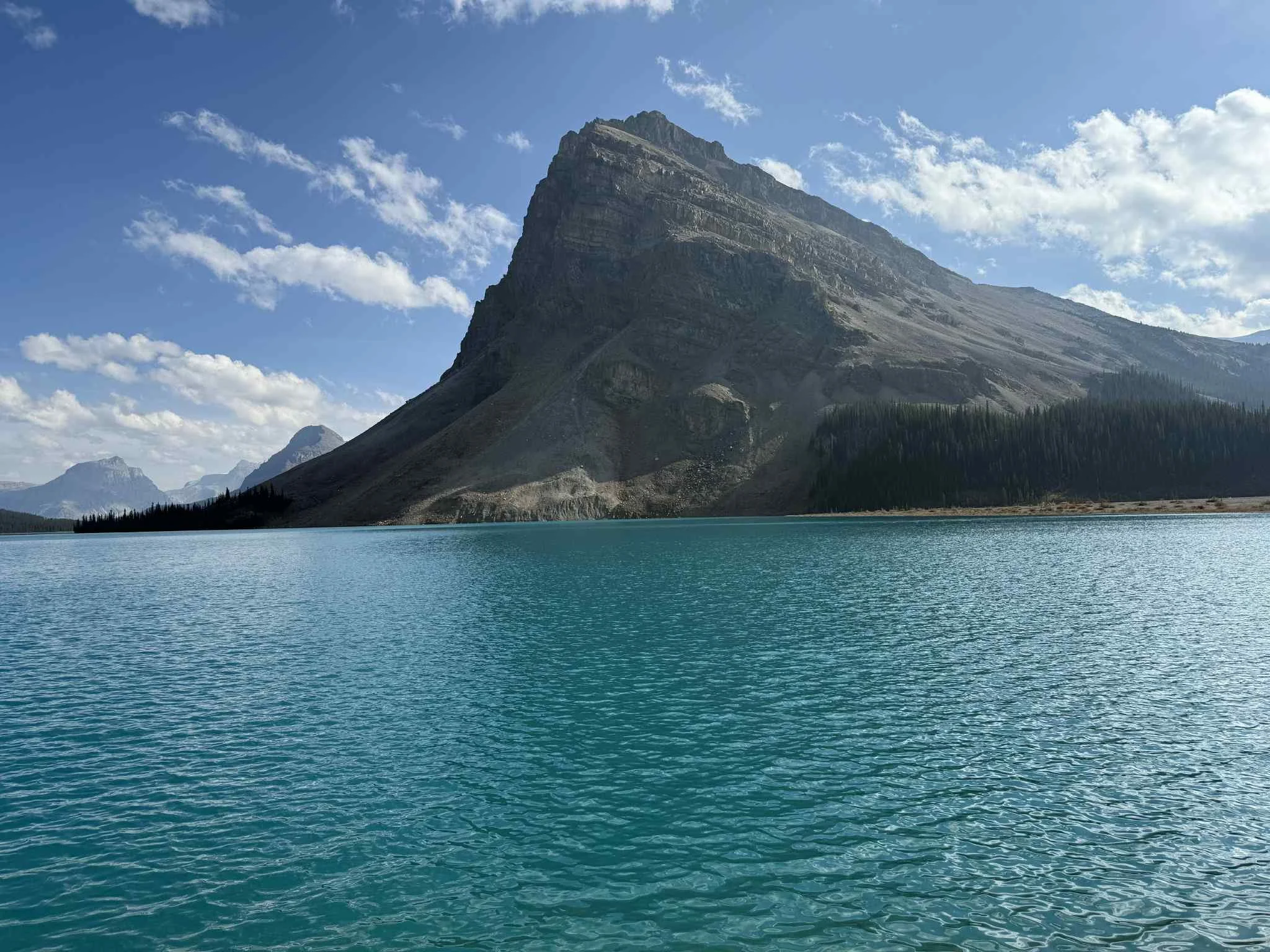 A large mountain with rocky slopes and a flat top, next to a serene turquoise lake, under a partly cloudy sky.
