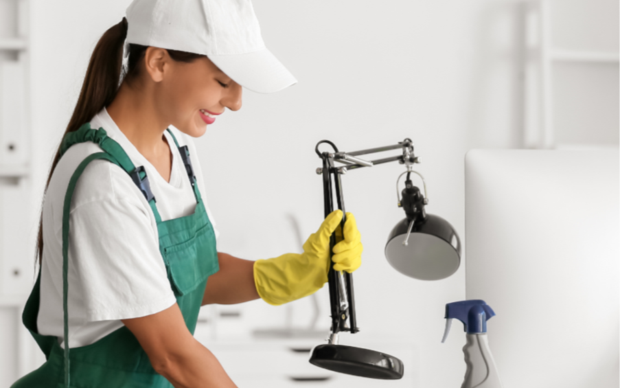 A woman dressed in a white cap, white shirt, green apron, and yellow rubber gloves holding a desk lamp in her hand, standing in a room with cleaning supplies.