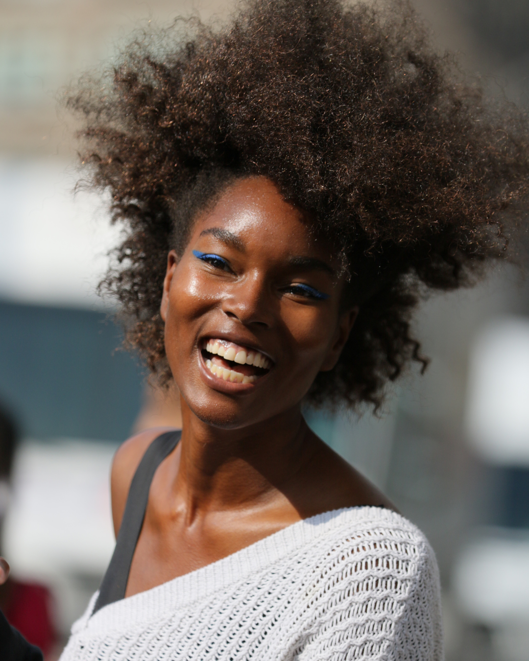 A woman smiling, with curly hair and blue eye makeup, outdoors in bright sunlight.