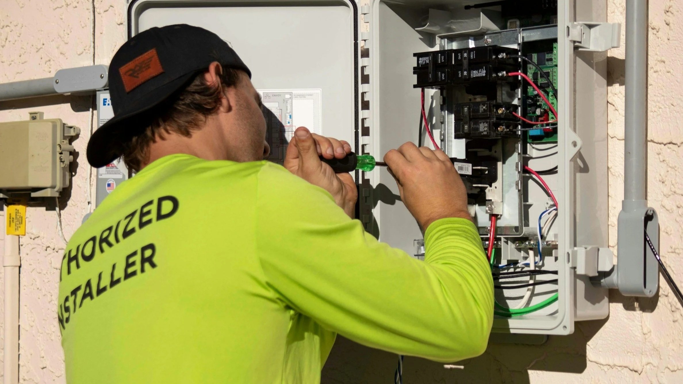 A worker in a neon yellow shirt and black baseball cap is using a screwdriver to work on an open electrical panel mounted on a beige wall.