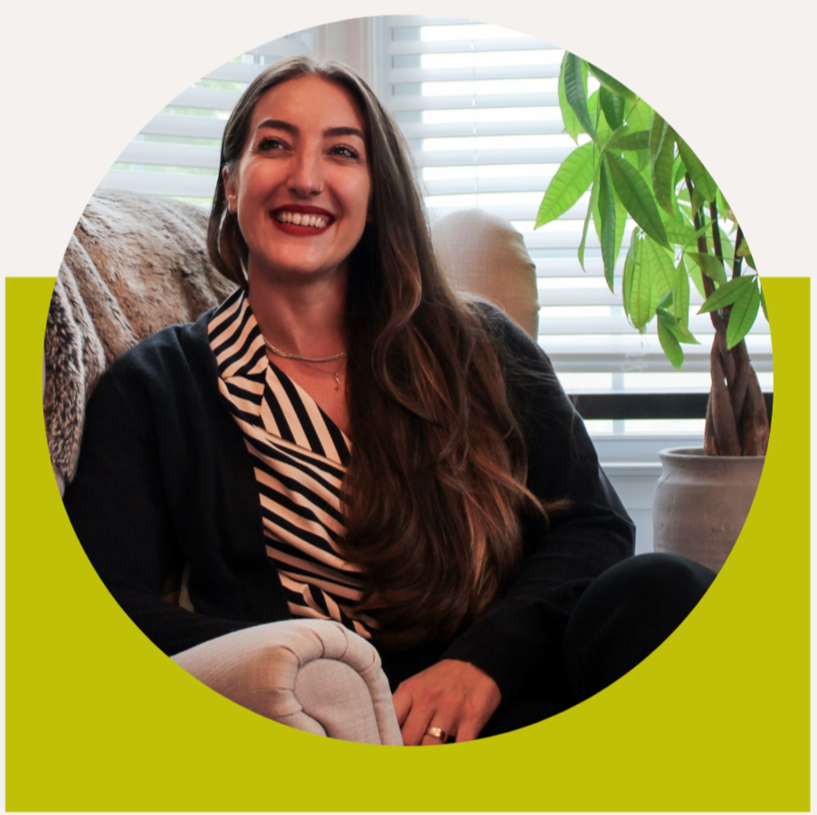 A woman with long brown hair smiling, sitting on a beige armchair in a room with white blinds and a green houseplant.