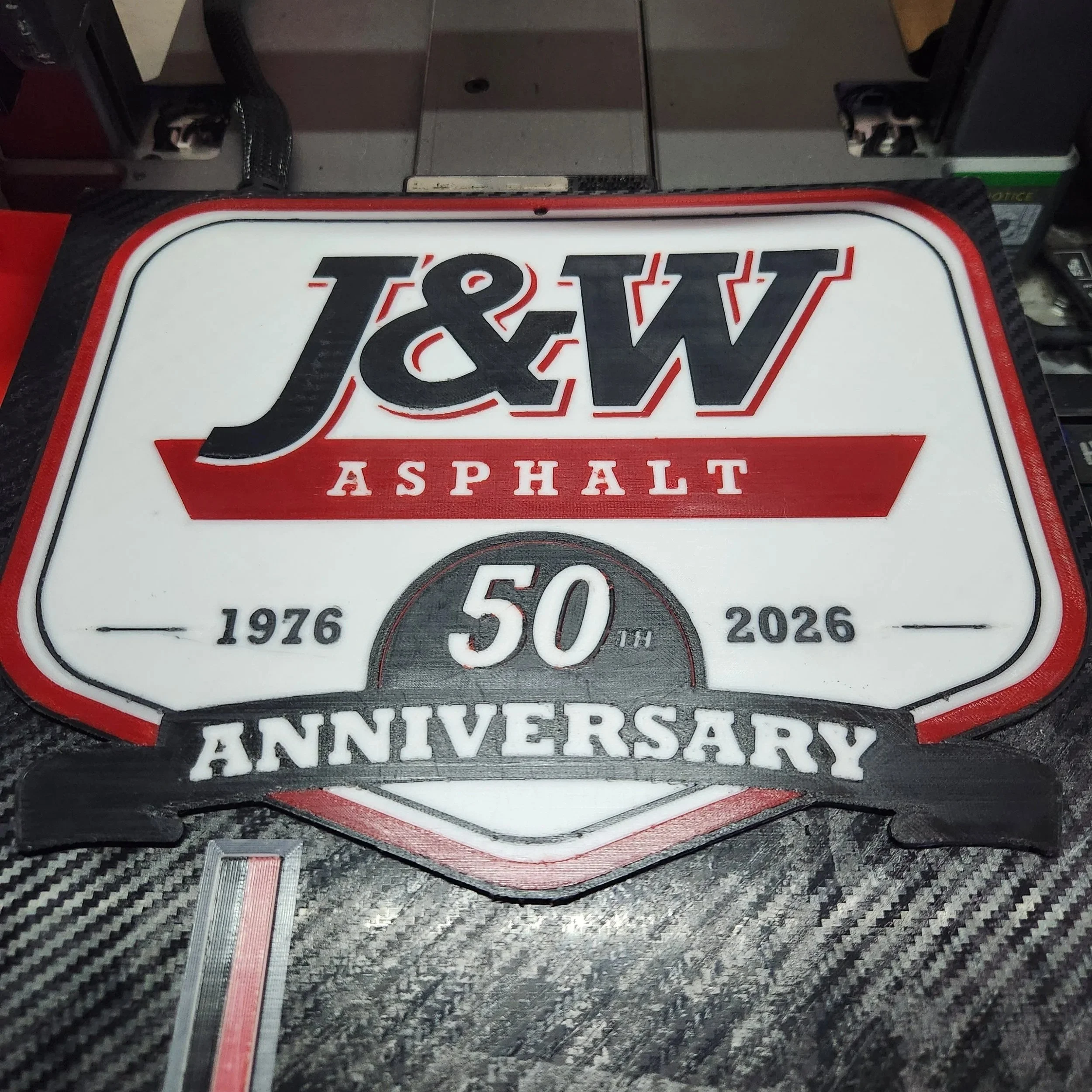 A decorative anniversary sign with the words '50th Anniversary' and a vintage-style design, placed on a textured surface with a carbon fiber pattern.